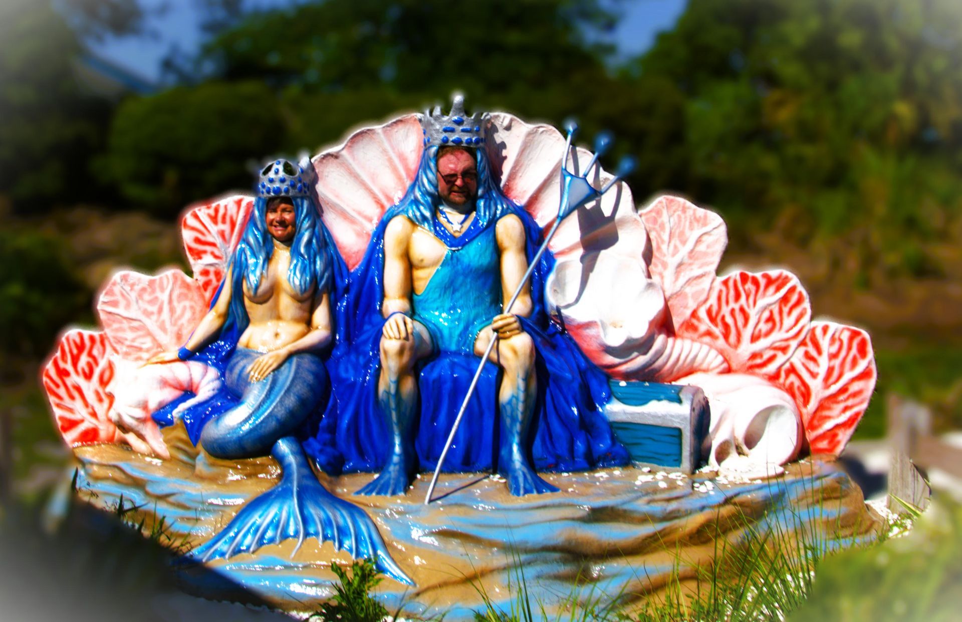 Sculpture of a sea king and mermaid seated on a seashell throne, blue and pink, outdoor setting.