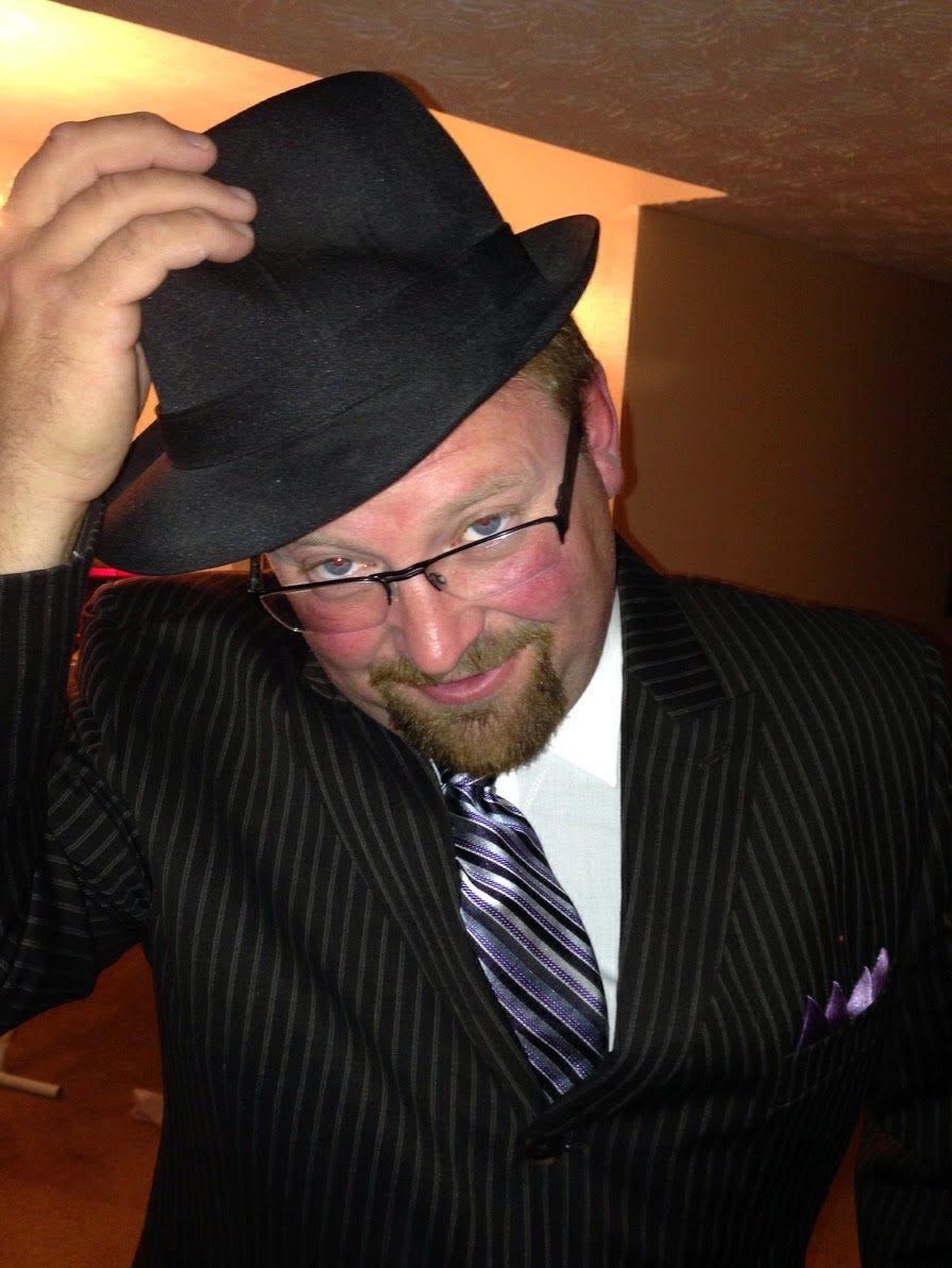 Man in pinstripe suit, fedora, and glasses tips hat, smiling, indoor setting.