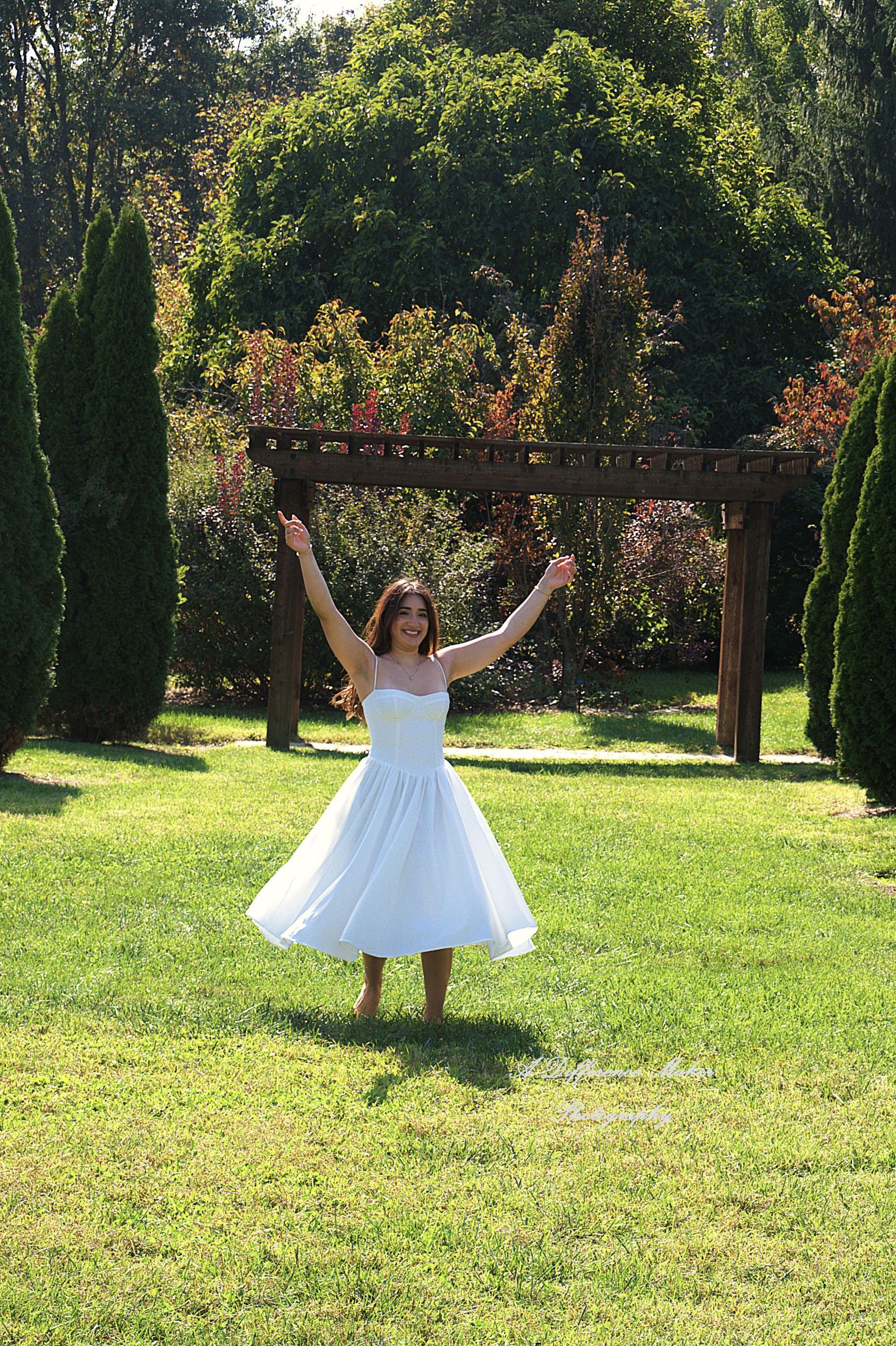 Woman in white dress with arms raised, standing in grassy yard near a wooden pergola.