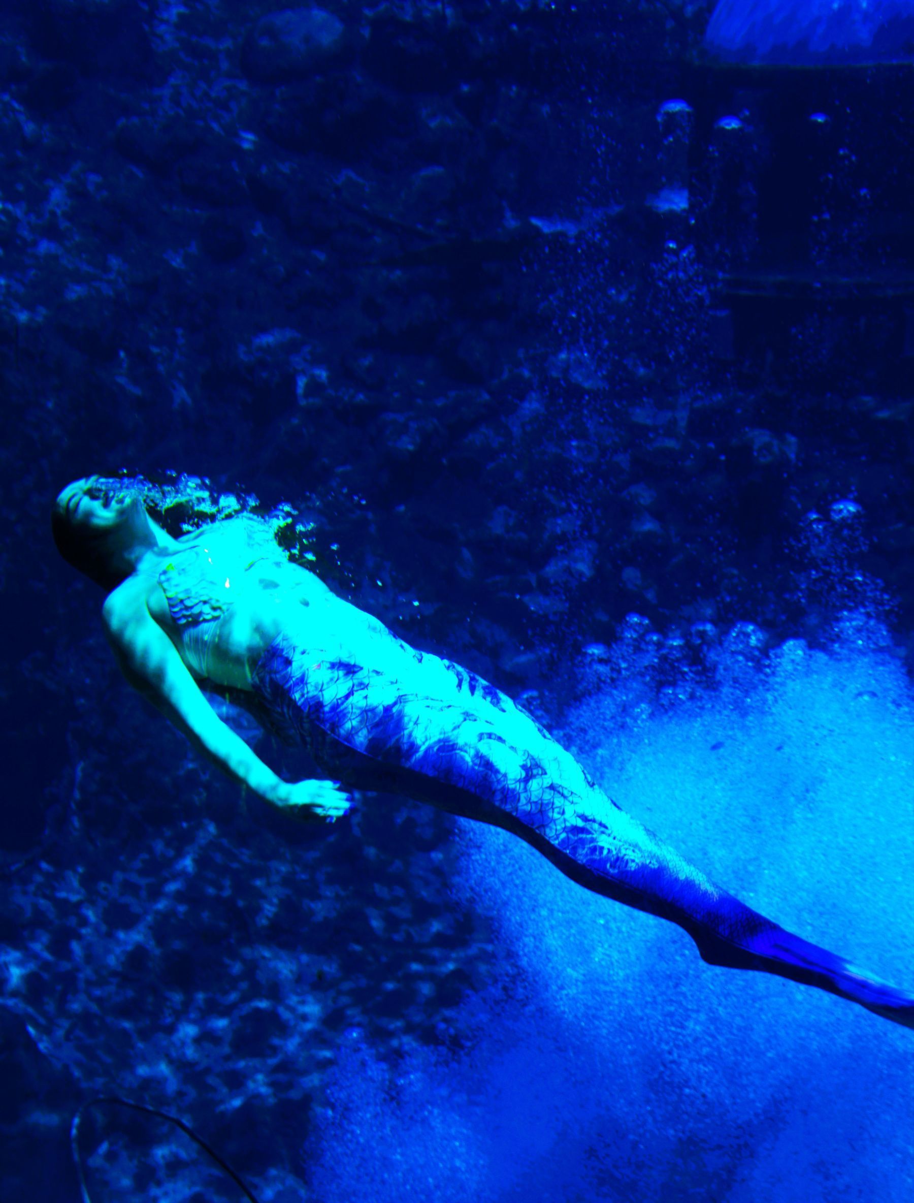Mermaid swims underwater in a blue environment; the tail is blue, the body is fair-skinned.