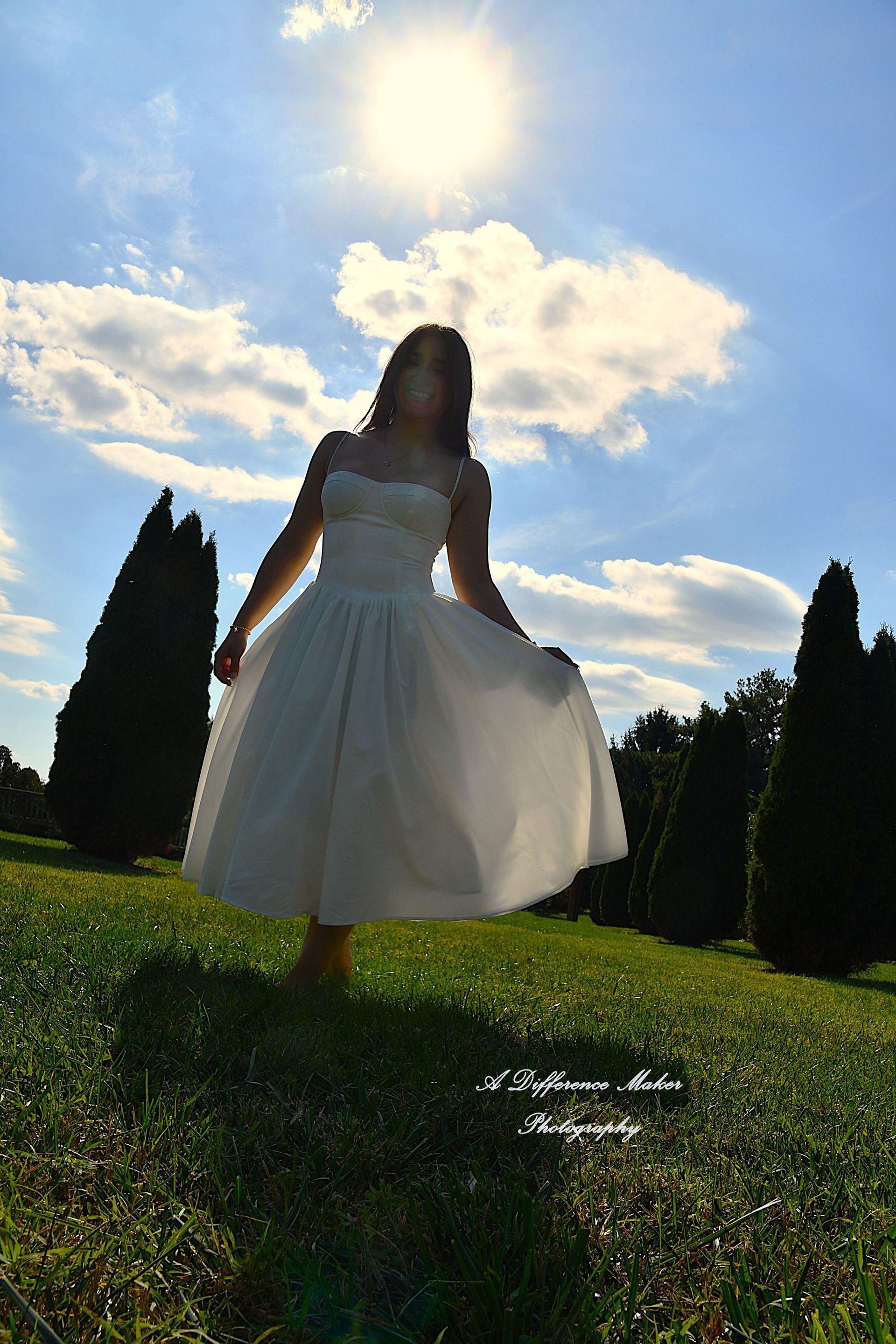 Woman in a white dress stands in a sunny field, holding skirt. The sun shines brightly with blue sky and trees.