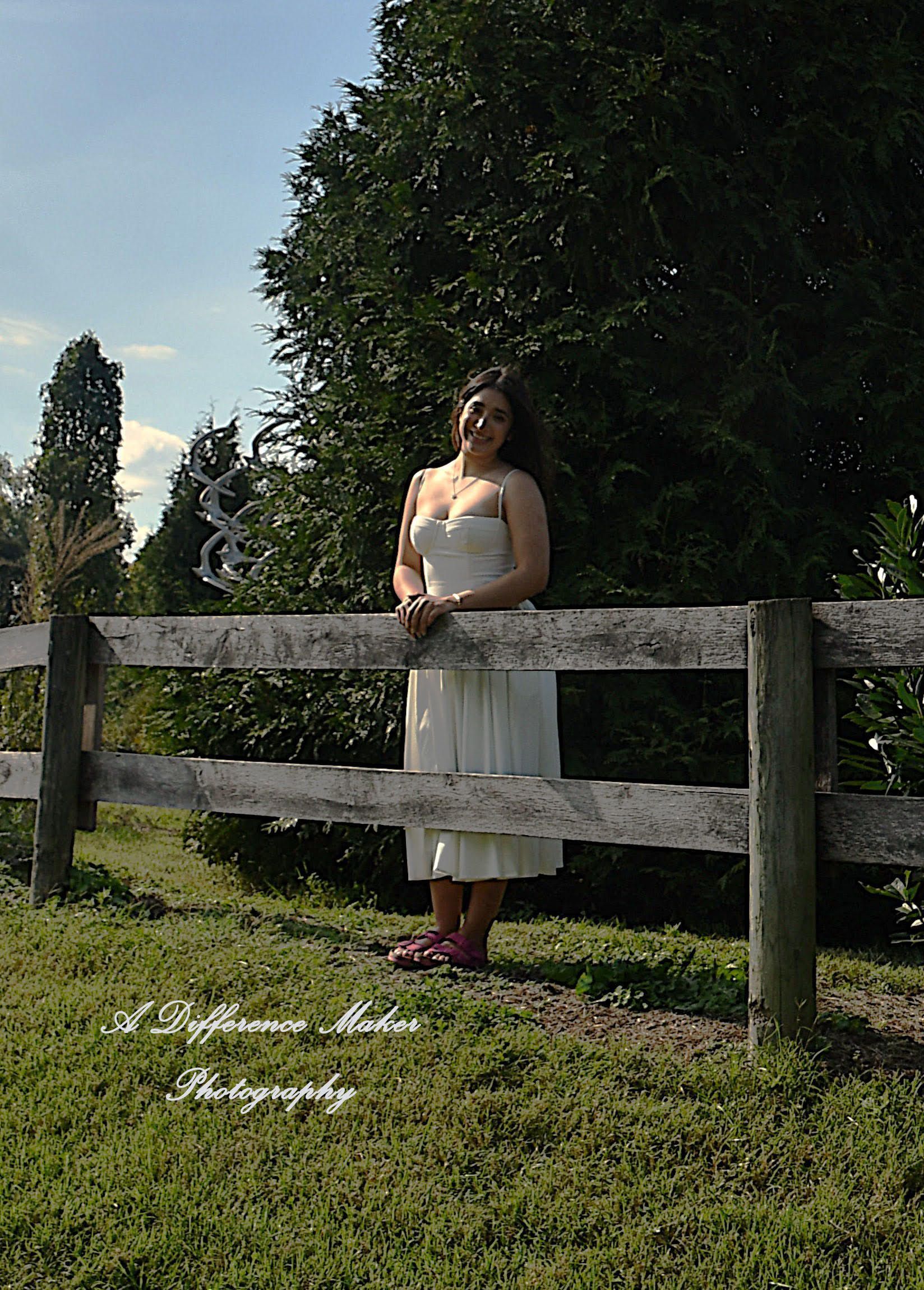 Woman in white dress standing by a white fence on a grassy field; trees in background.