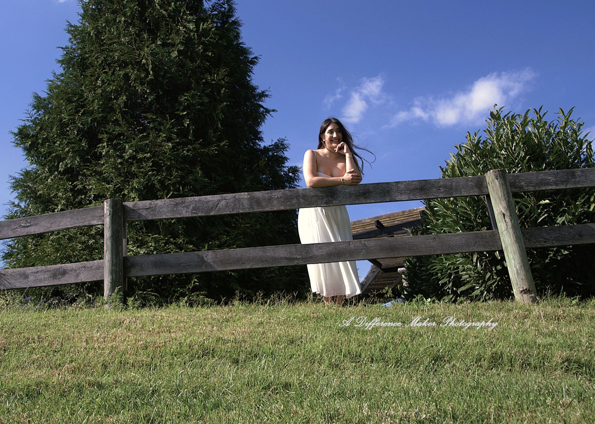 Woman leans on a wooden fence, smiling. Set in a grassy field, with trees and a blue sky in the background.