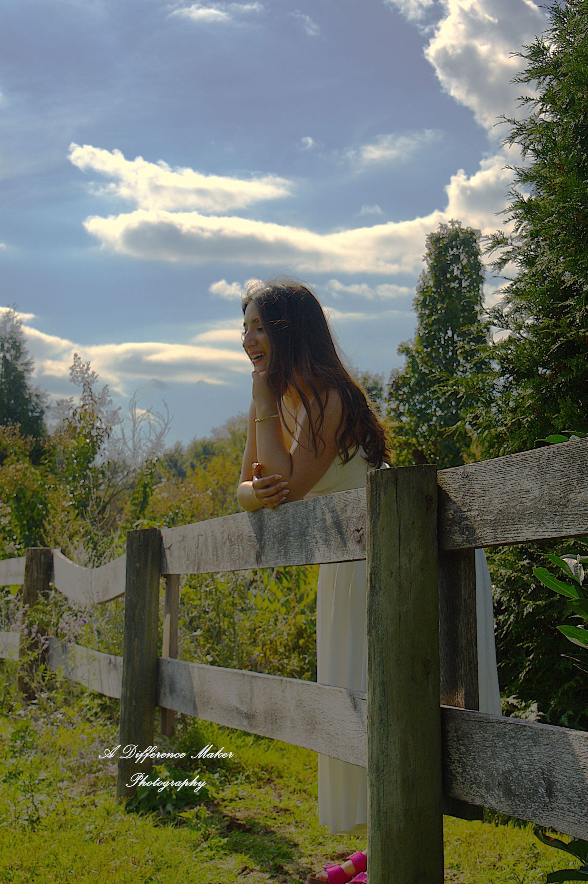 Woman leaning on a wooden fence, looking towards a blue sky with clouds, surrounded by greenery.