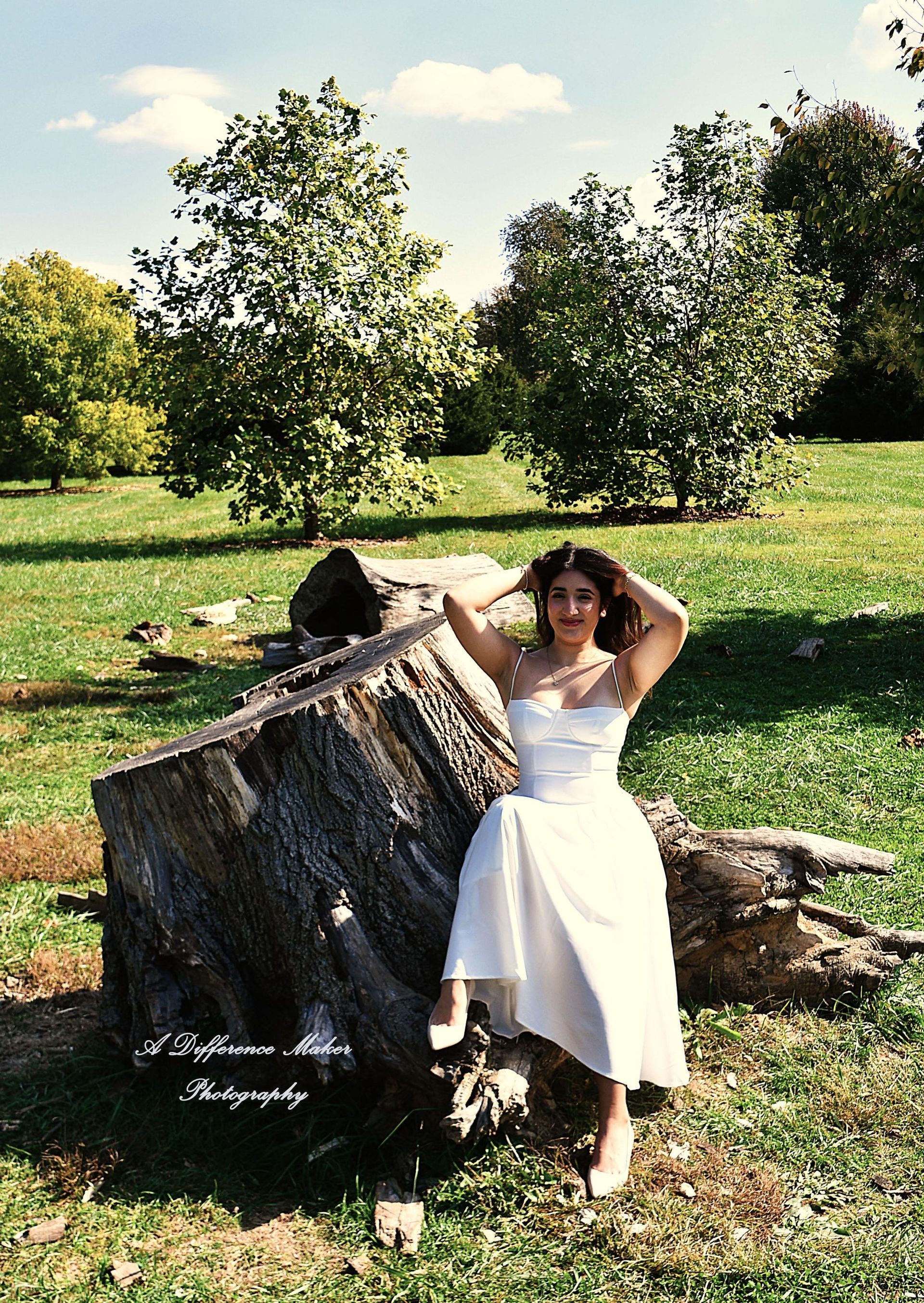 Woman in a white dress poses on a large log in a sunny park.