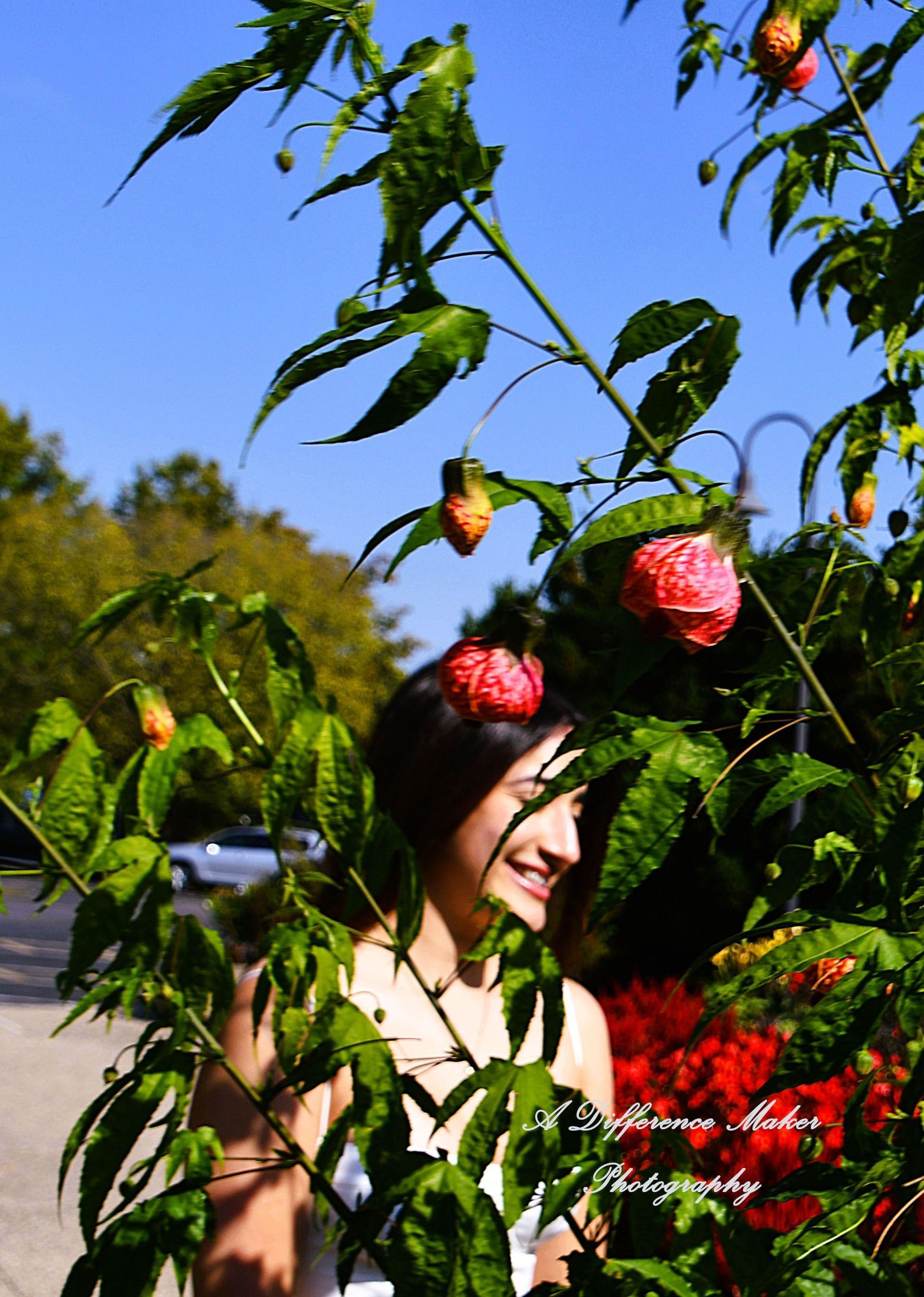 Woman smiling amongst vibrant red-orange flowers and green foliage, against a blue sky.