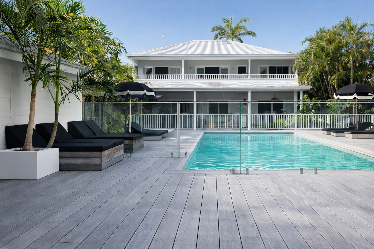 A white two-story house with a pool and wooden deck. Lounge chairs and umbrellas surround the pool.