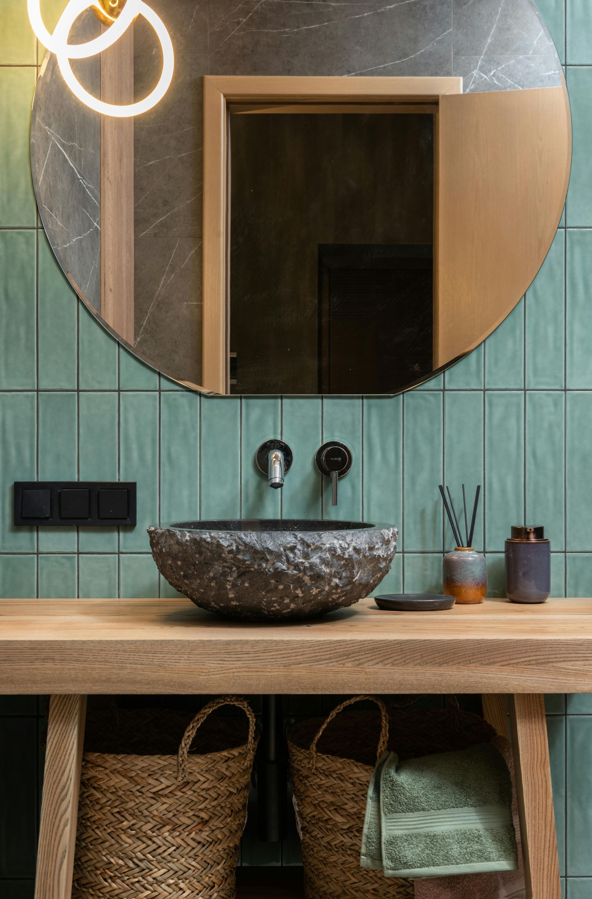 Bathroom with a round mirror, stone sink, and aqua tile backsplash above a wooden vanity. Wicker baskets underneath.