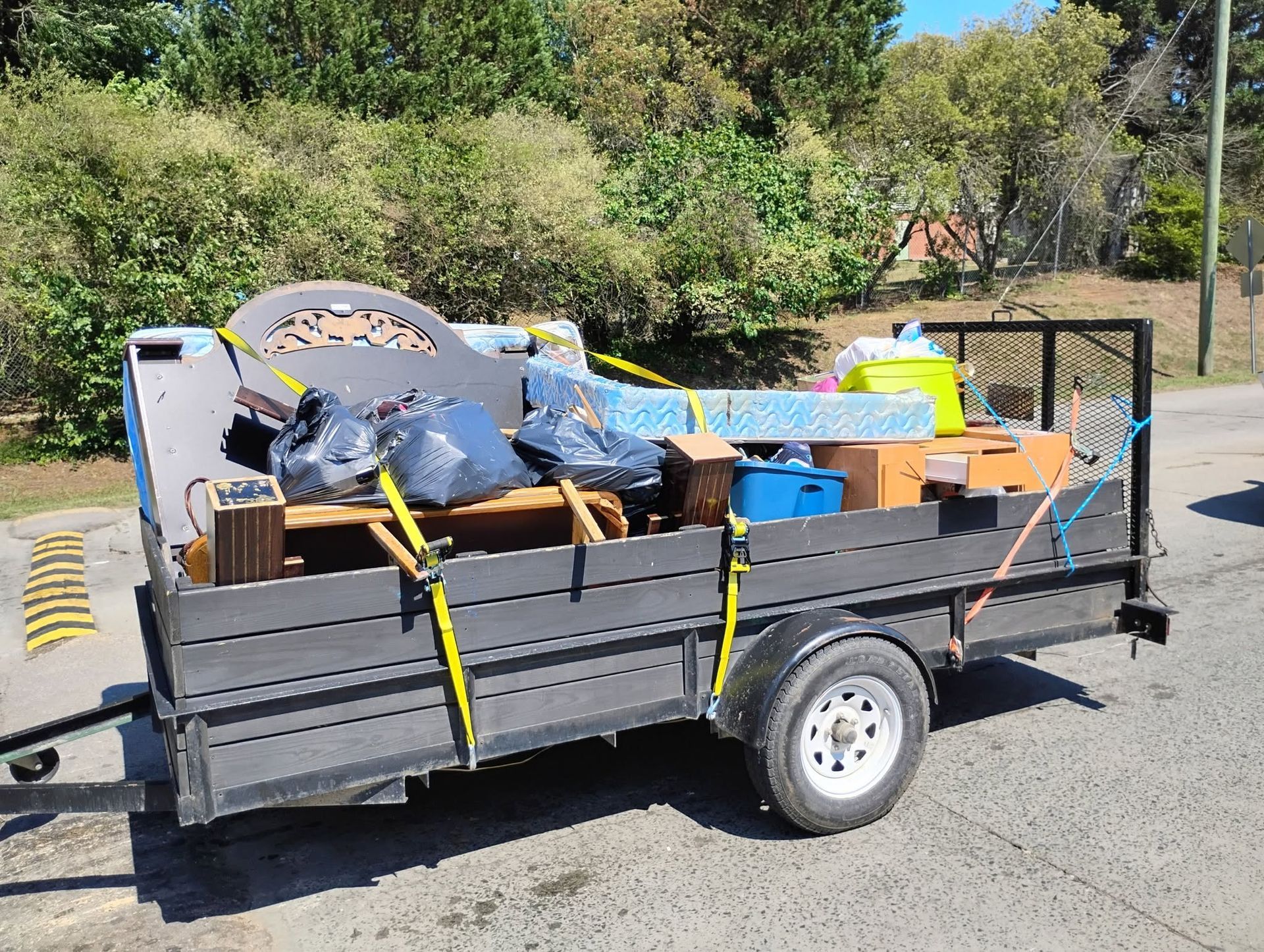 Gray utility trailer piled with assorted moving boxes and furniture on a roadside, with trees in the background