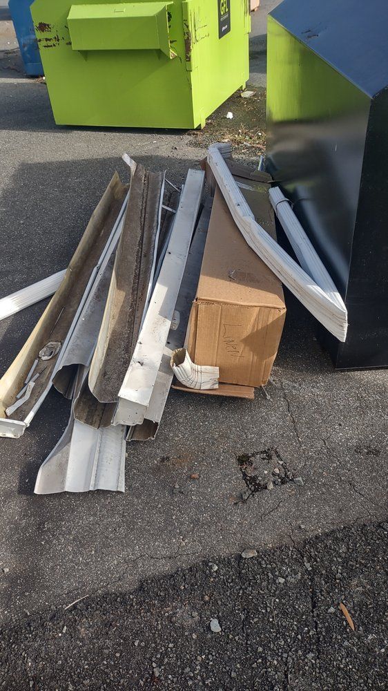 Pile of broken metal and cardboard debris on pavement beside green dumpsters.