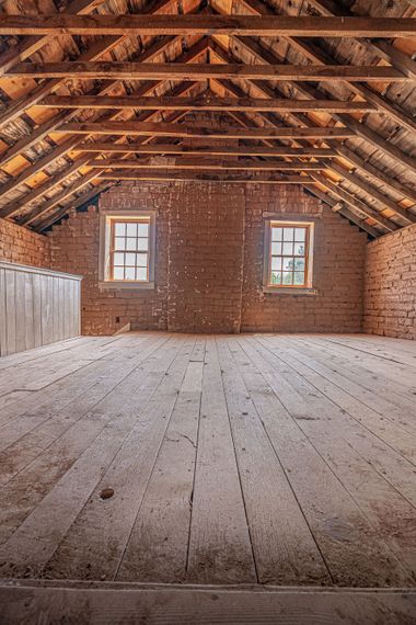 Empty rustic attic with exposed wooden beams, brick walls, and two small windows