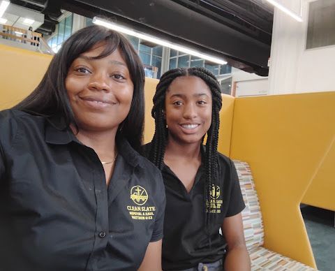 Two women in black collared shirts smiling indoors, seated beside a yellow wall.