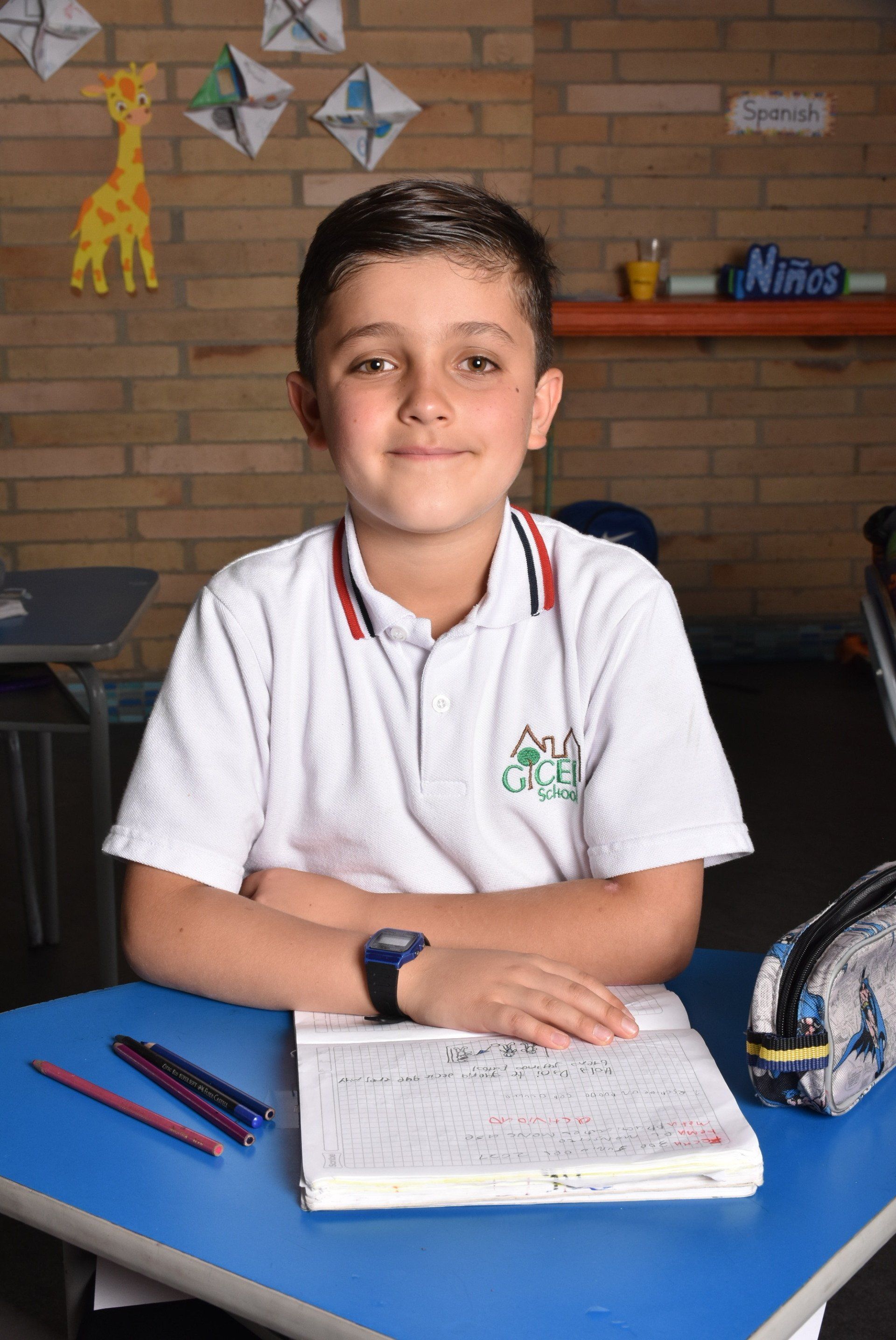 Una niña sentada en un pupitre azul con cuadernos y lápices en un aula, sonríe a la cámara.