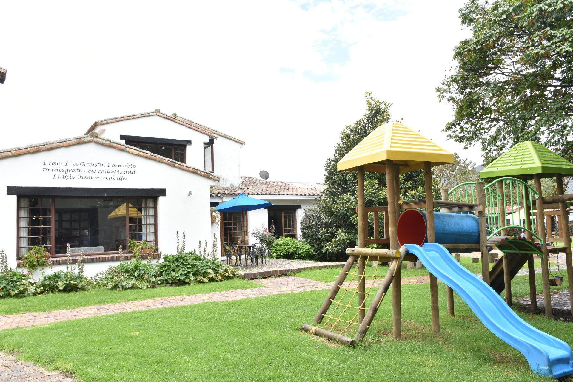 Parque infantil con tobogán azul y estructuras de juego con techo amarillo en un césped junto a un edificio blanco.