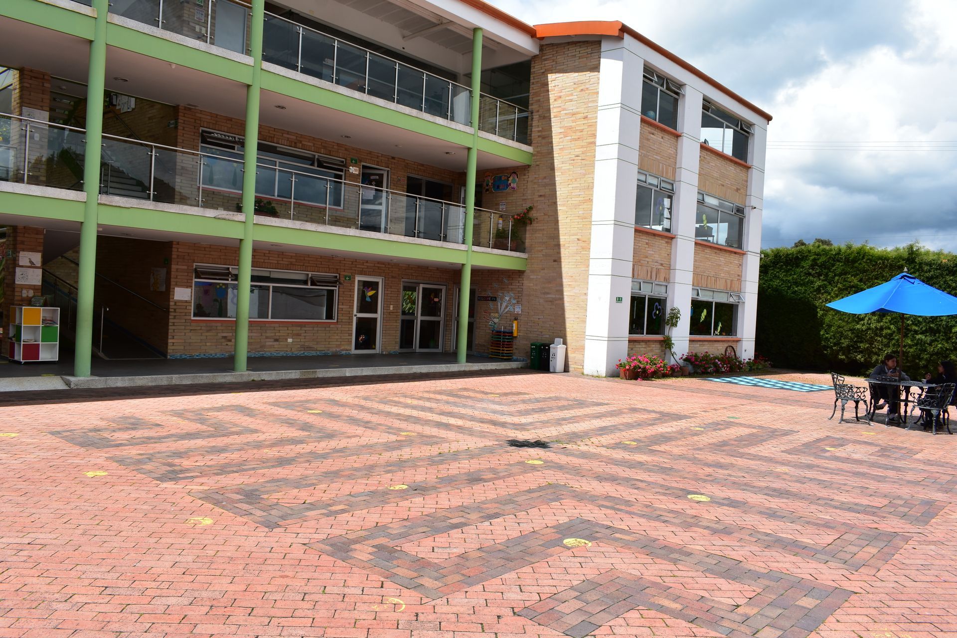 Patio del campus con un edificio de varias plantas, suelo de ladrillo y sombrillas azules a la derecha.