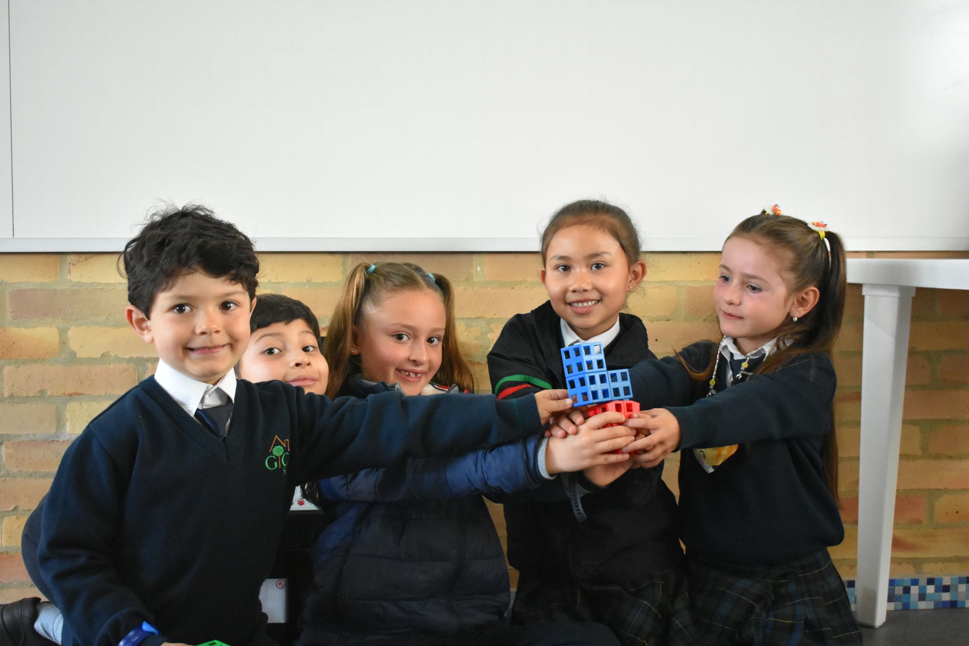 Cinco niños con uniformes oscuros sonríen mientras sostienen juntos una torre de bloques azules y rojos en un aula.
