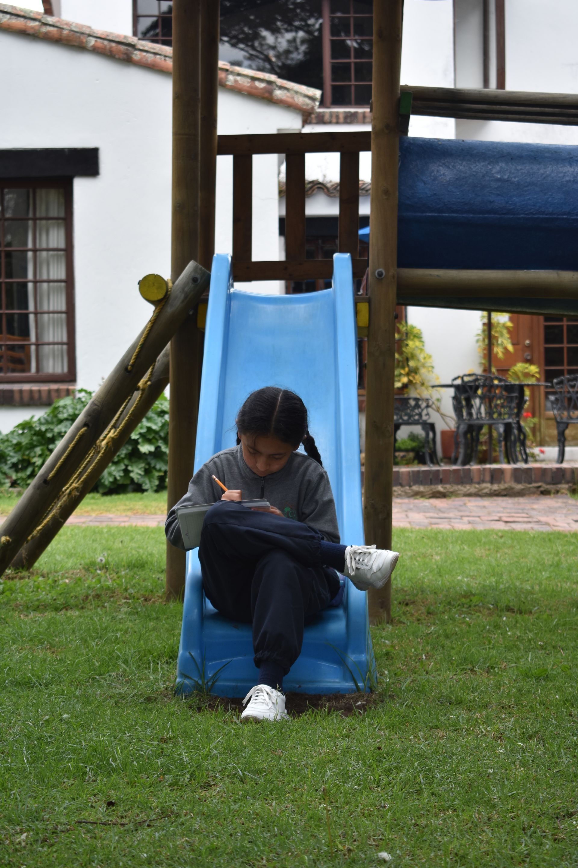 Una persona sentada en un tobogán azul, mirando una tableta en un parque infantil cubierto de césped.