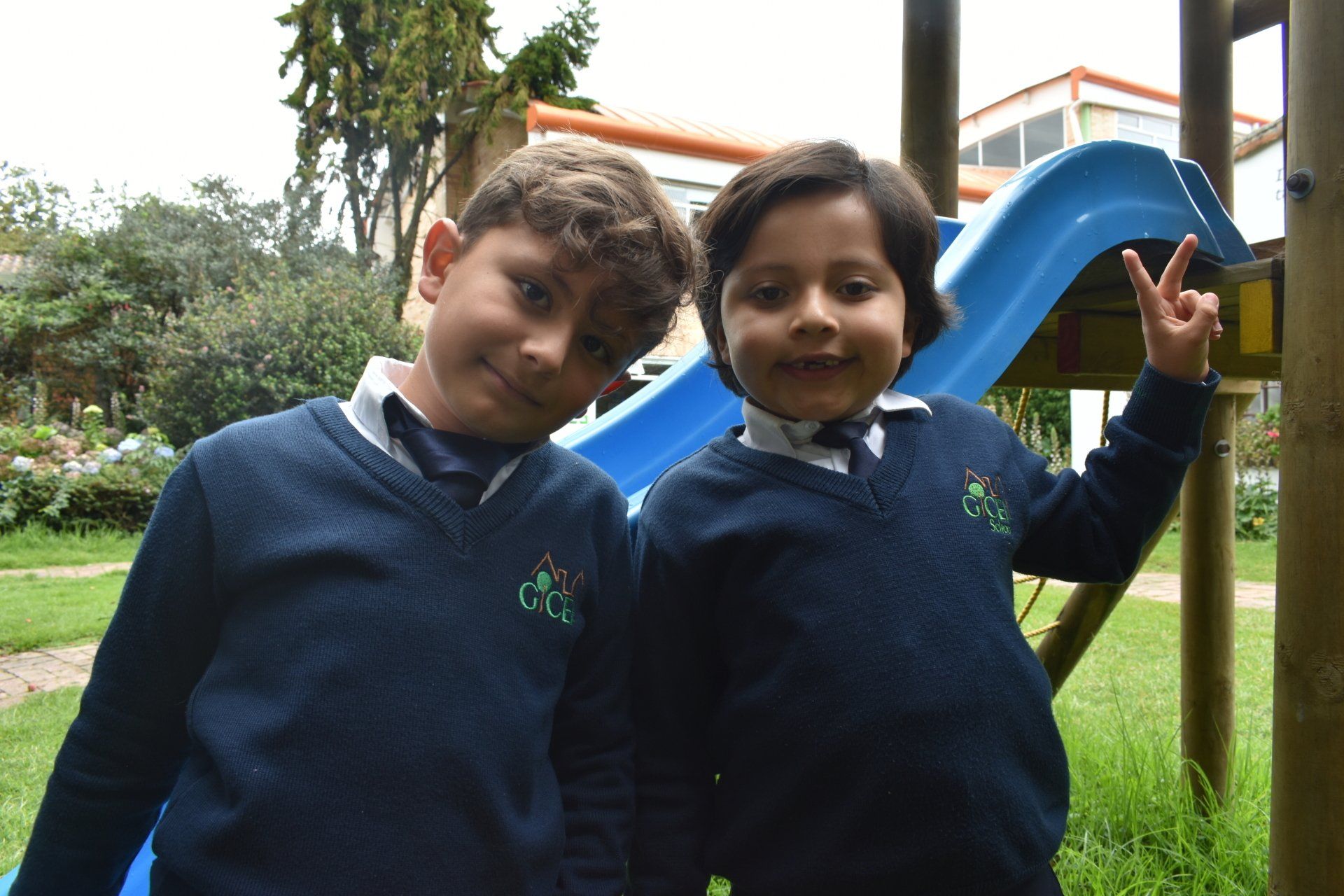 Dos niños con suéteres azul marino posan sonriendo cerca de un tobogán en un parque infantil; uno de ellos hace el signo de la paz.
