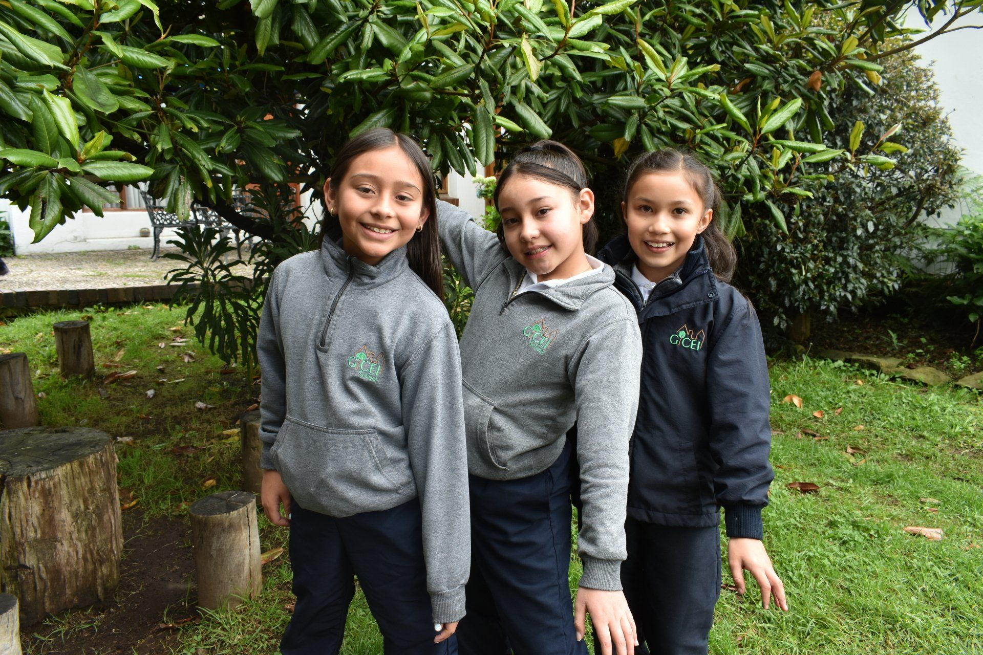 Tres niños de pie al aire libre junto a un árbol frondoso, vestidos con chaquetas grises y oscuras, sonriendo a la cámara.