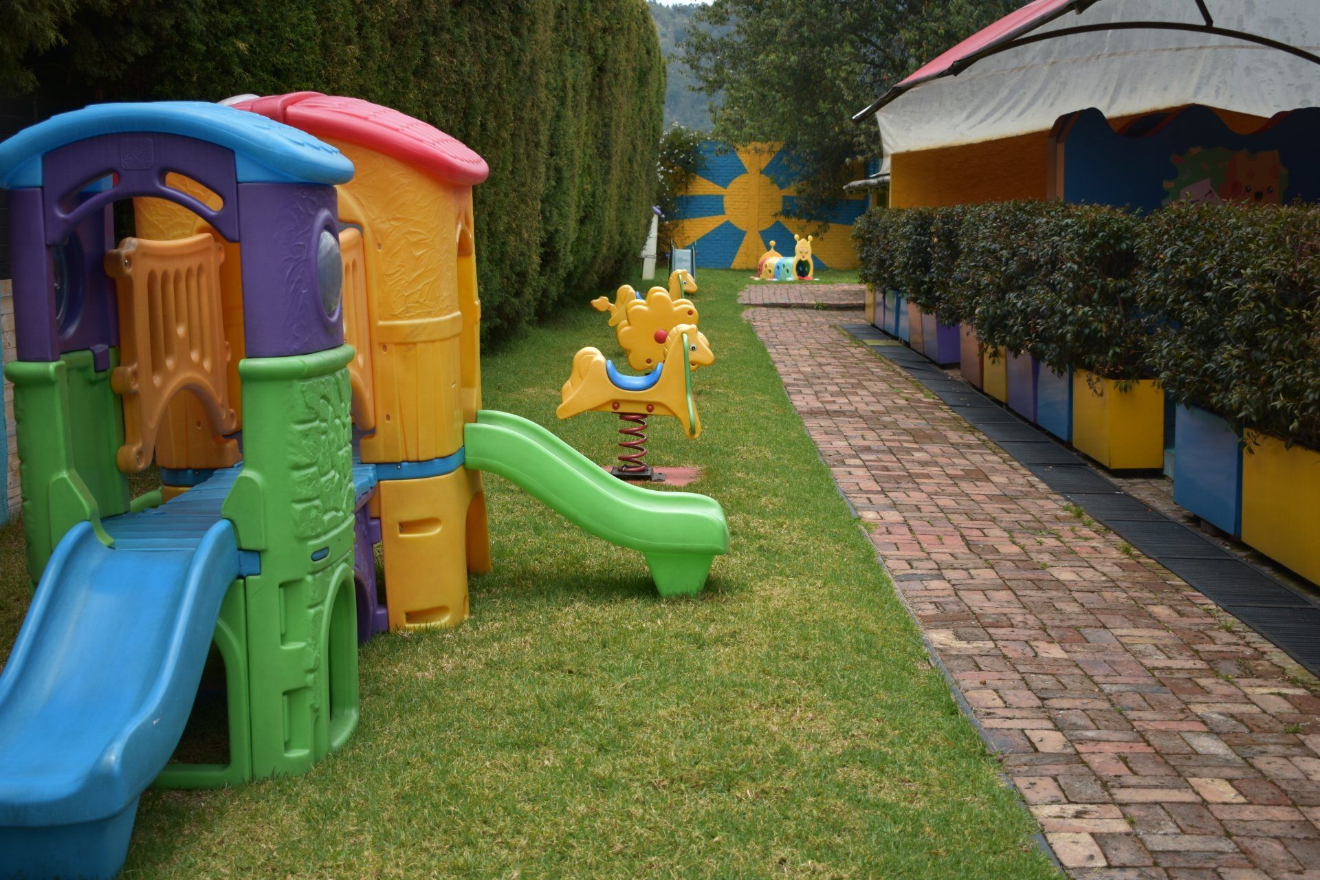 Parque infantil colorido con toboganes y casita de juegos junto a un sendero de piedra y un patio bordeado de setos.