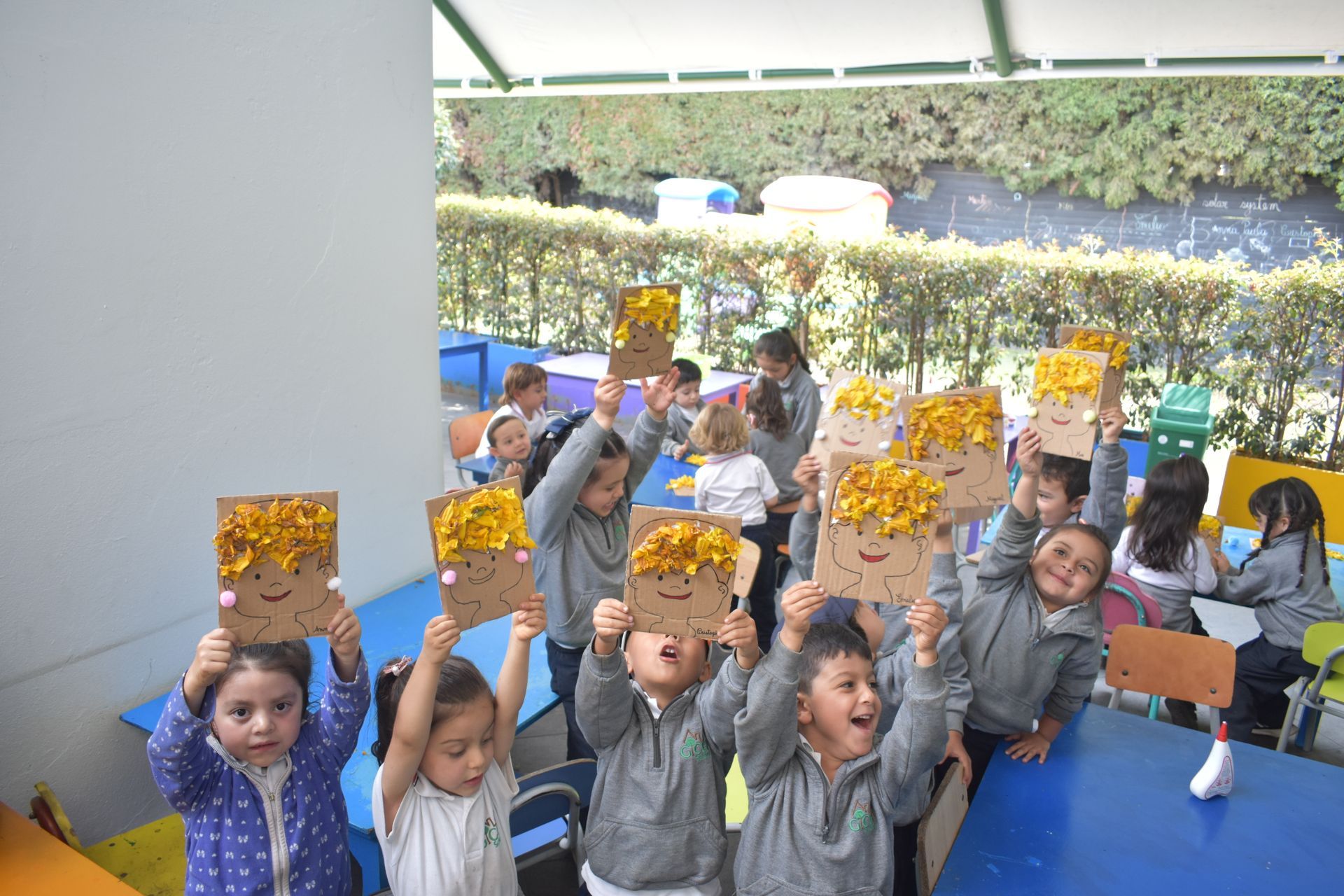 En el luminoso patio de un aula, niños con uniformes grises sostienen hojas de papel amarillas para dibujar.