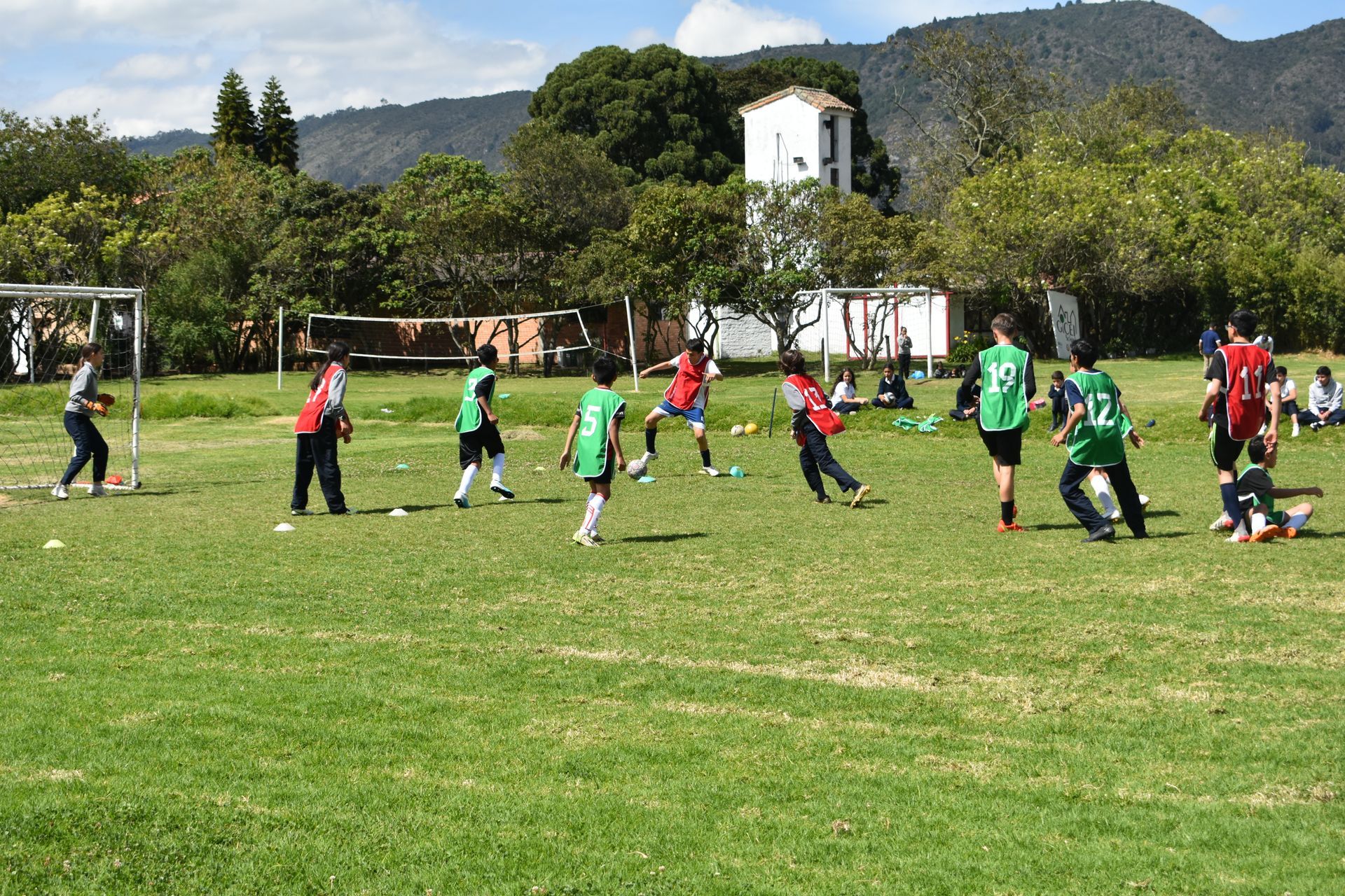 Niños jugando al fútbol en un campo de hierba, con montañas y un edificio blanco al fondo.