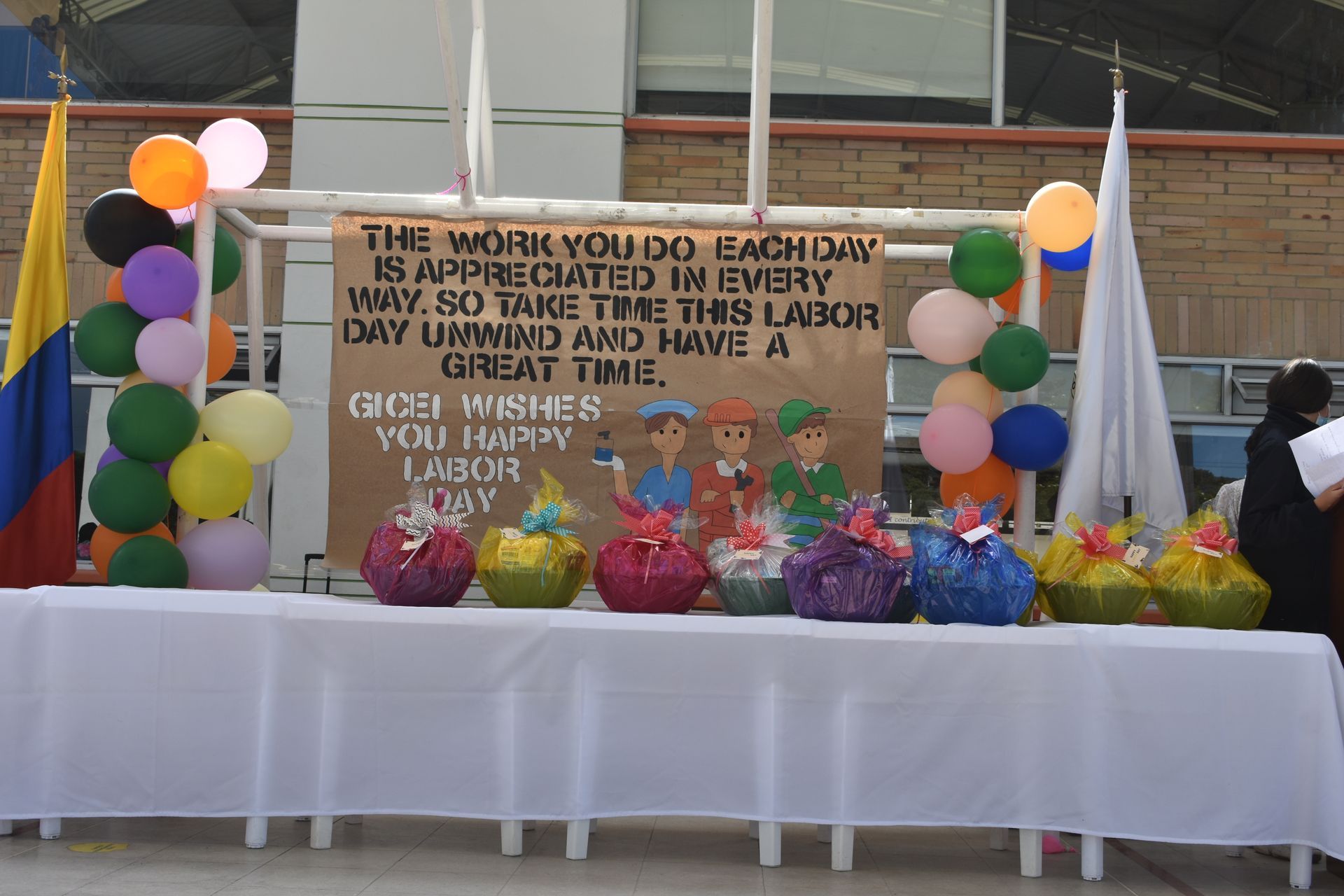 Mesa exterior decorada con globos de colores, peluches y una pancarta de agradecimiento en un escenario escolar.