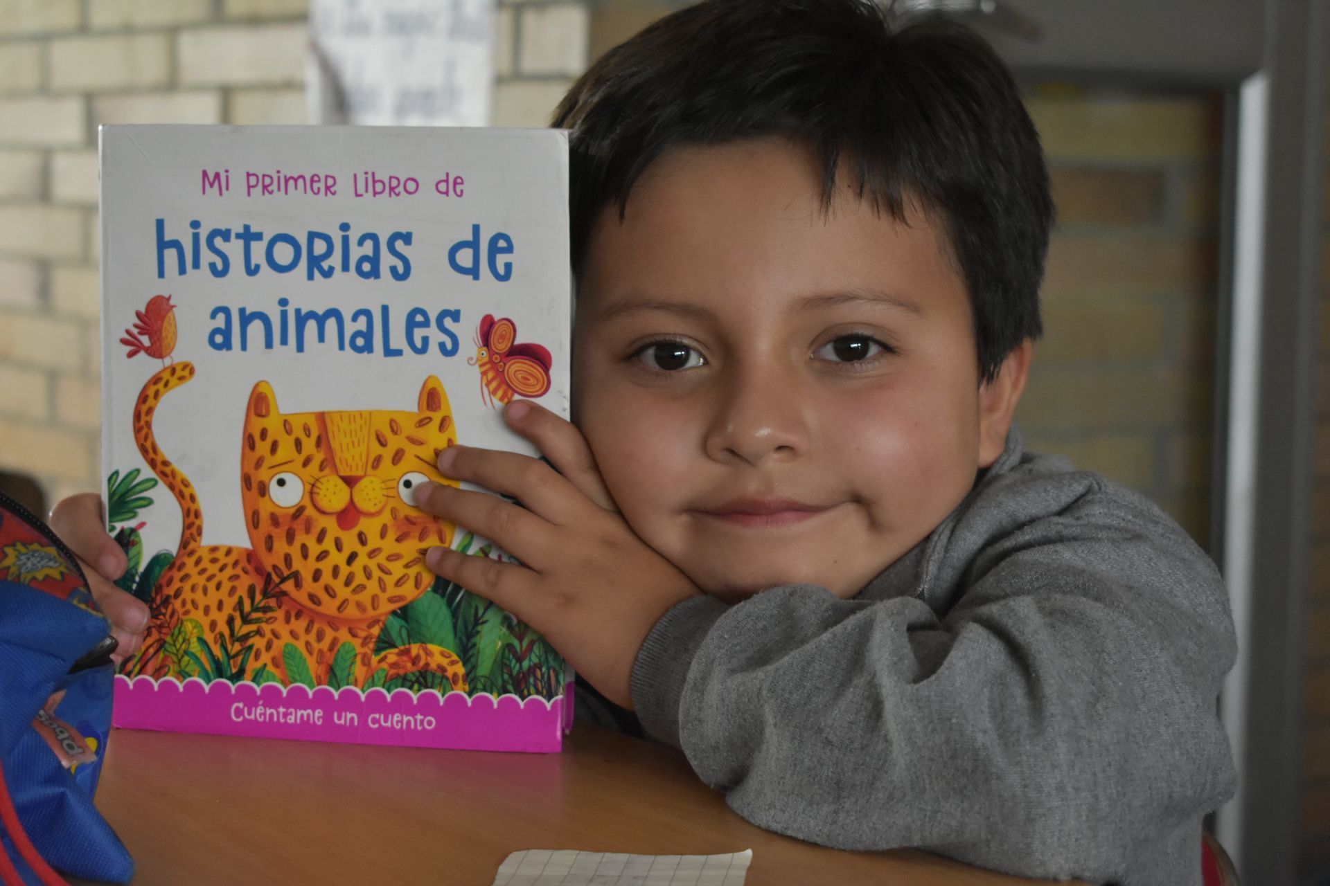 Un niño sostiene un colorido libro de “historias de animales” y sonríe sentado a una mesa.
