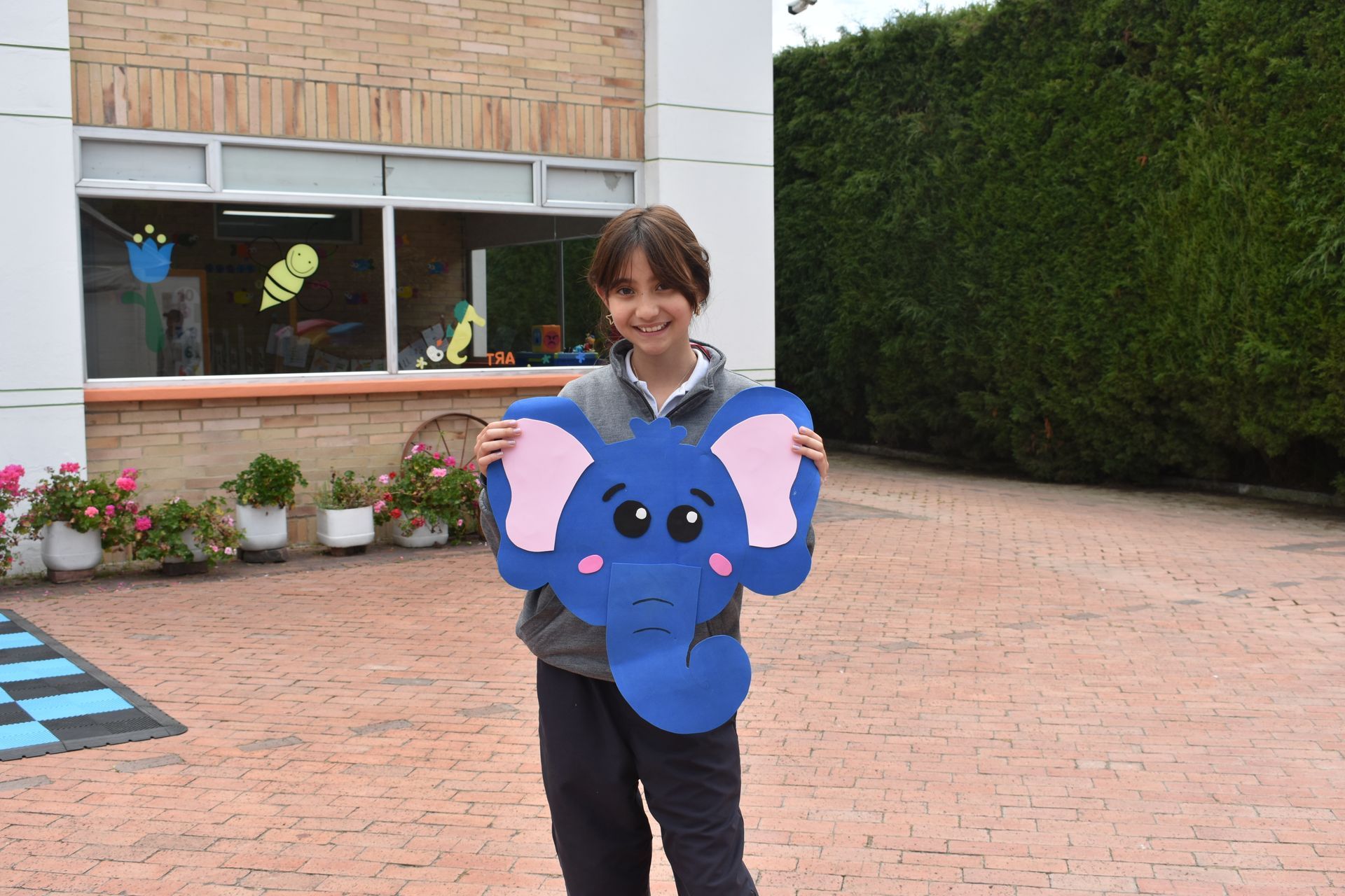 Persona sosteniendo una manualidad de elefante azul al aire libre en un patio, con orejas rosas y una cara sonriente.