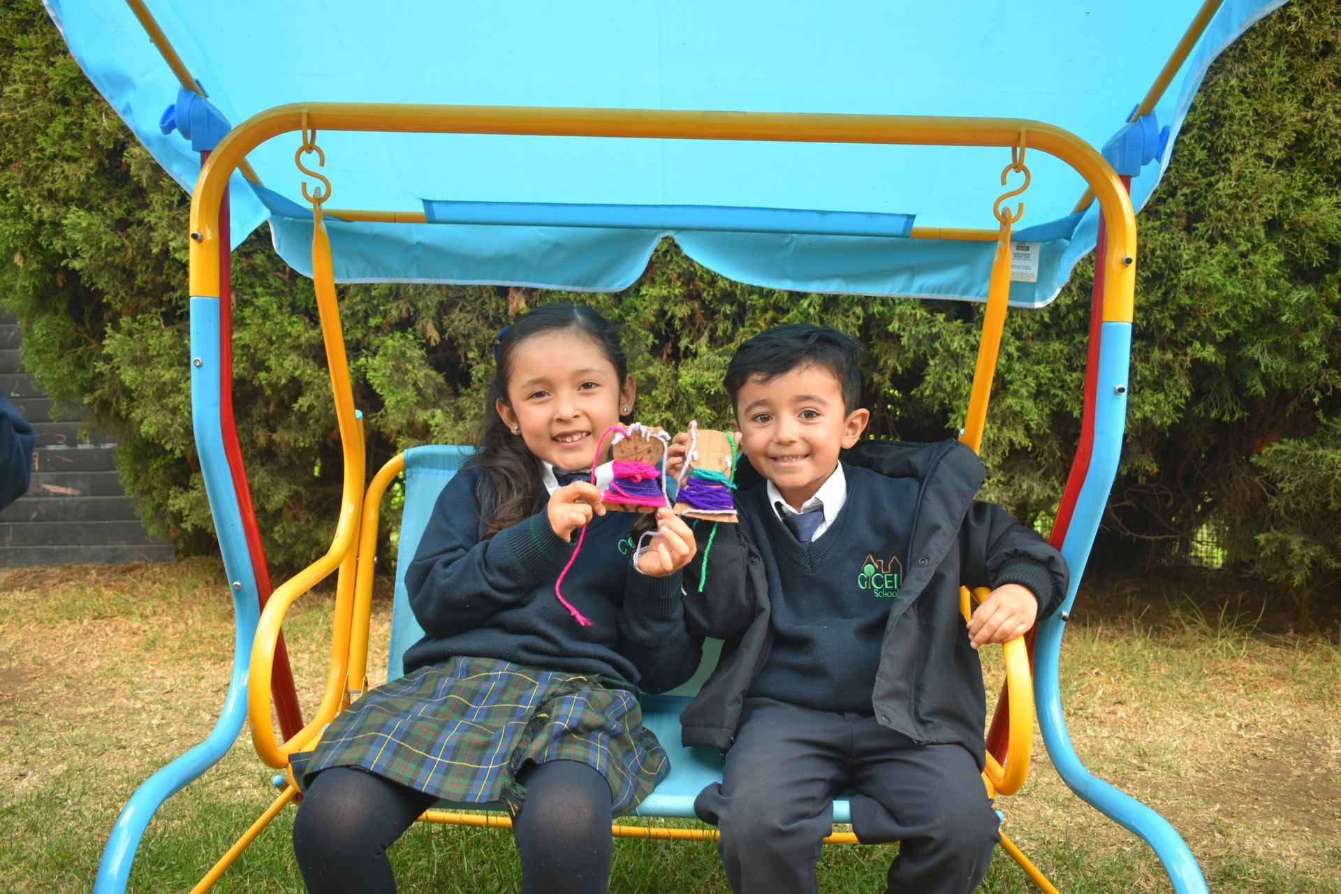 Dos niños están sentados sonriendo en un columpio azul y amarillo en un parque infantil al aire libre, sosteniendo un juguete.