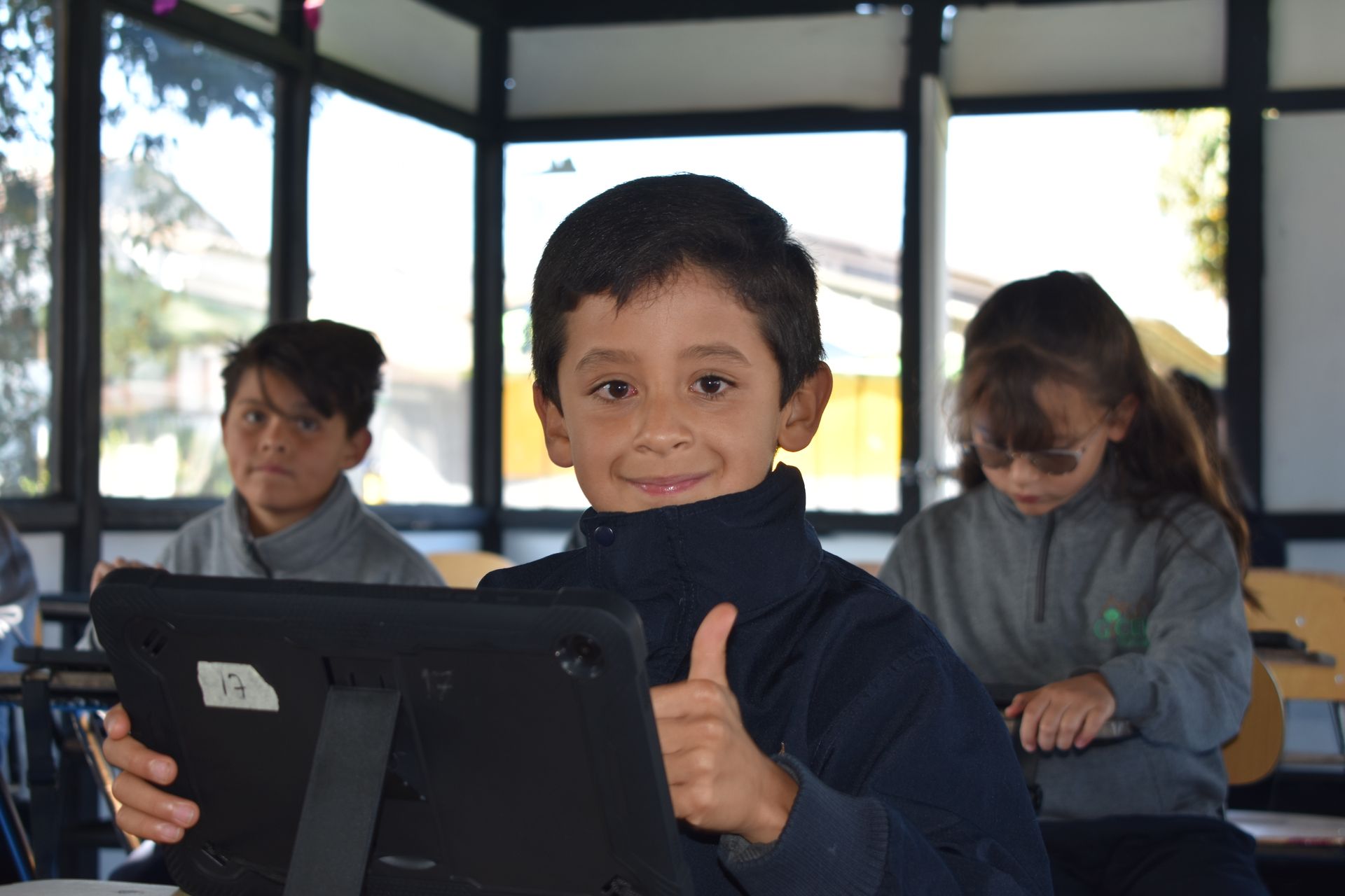 Un niño sonríe y levanta el pulgar mientras usa una tableta en un aula, con dos compañeros de clase al fondo.
