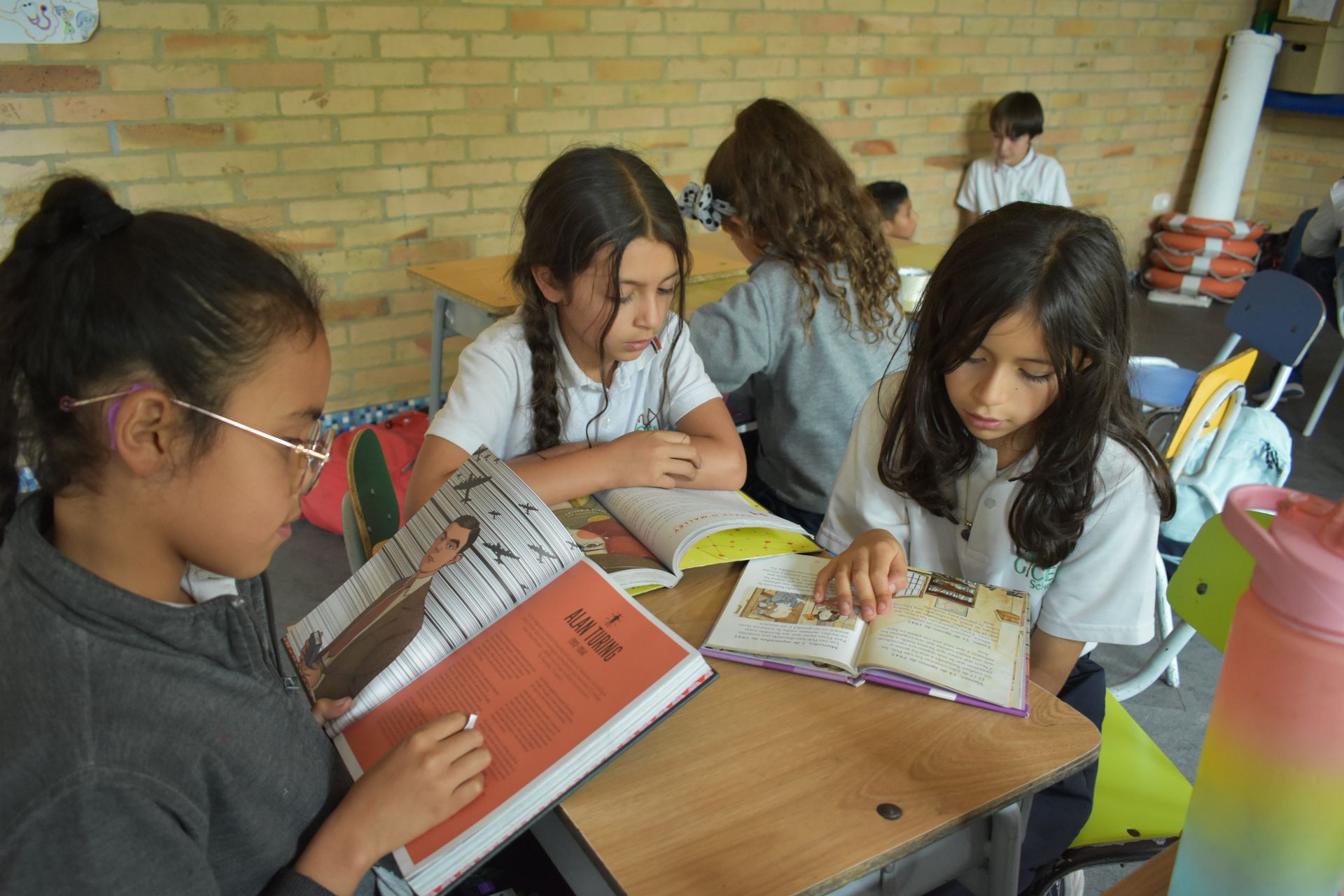 Niños leyendo juntos en una mesa en un aula, con libros y papeles abiertos.