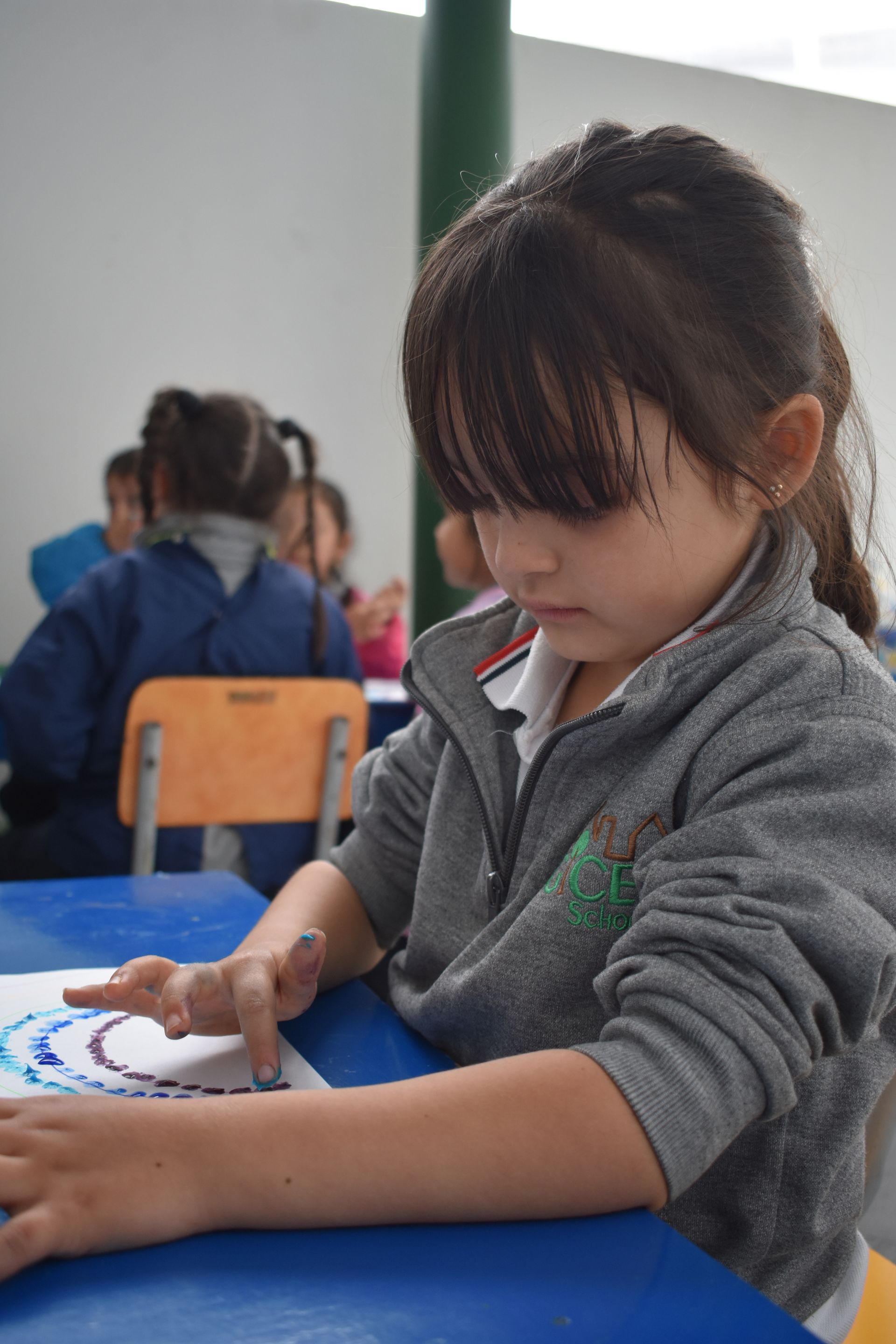 Un niño dibuja con pintura blanca en una mesa azul en un aula.