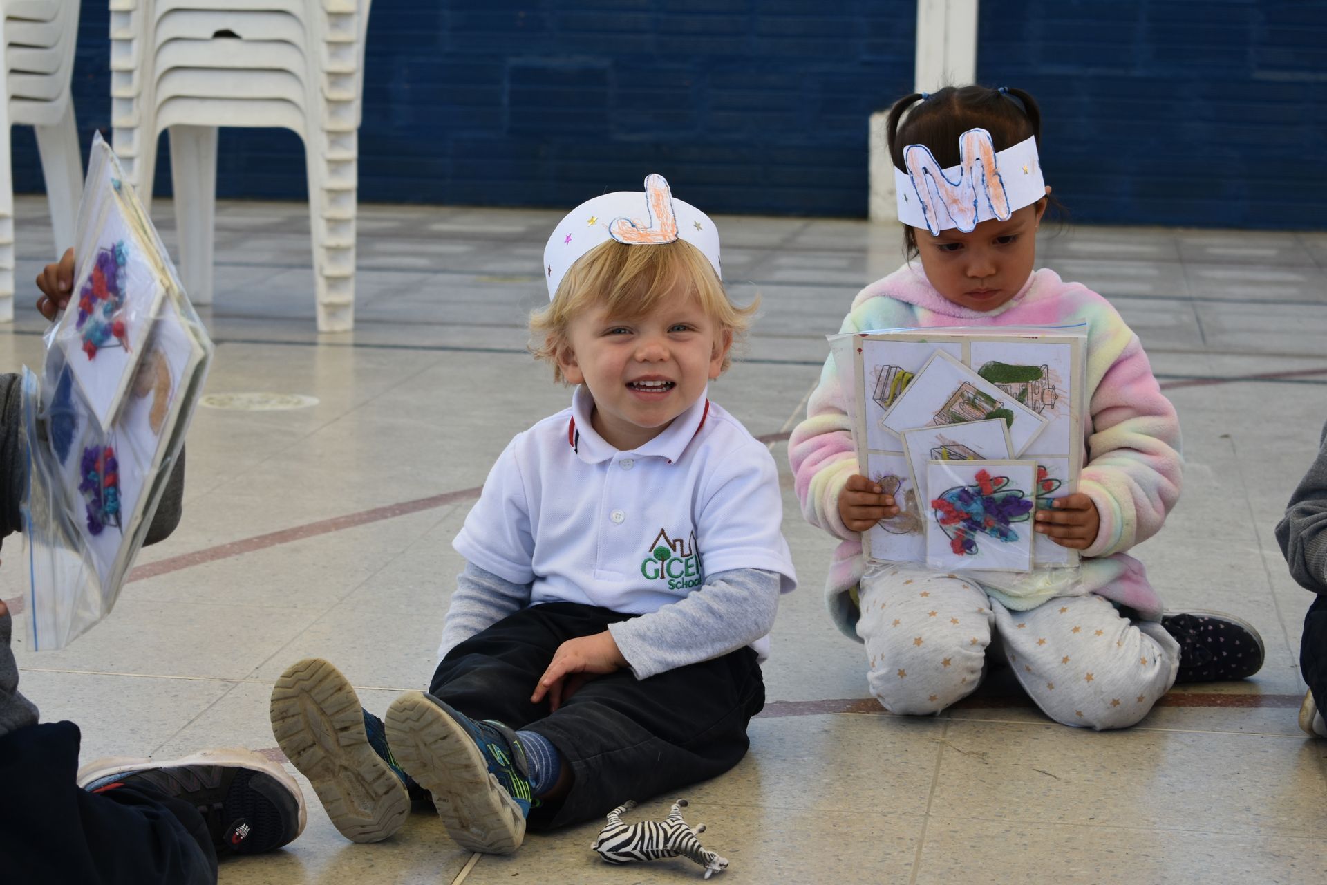 Dos niños sentados en el suelo de un gimnasio, sonriendo y sosteniendo papeles, con coronas de papel blancas y ropa escolar.