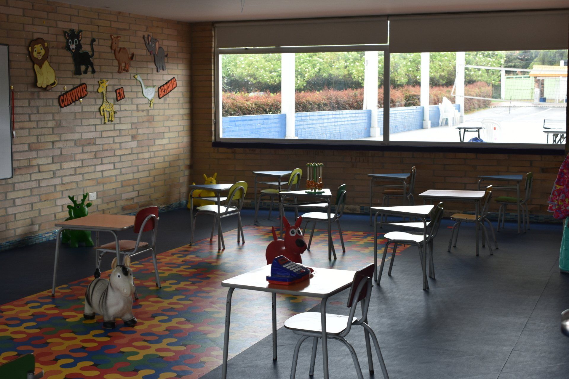 Aula vacía con mesas pequeñas, sillas coloridas y juguetes cerca de una gran ventana a la luz del día.