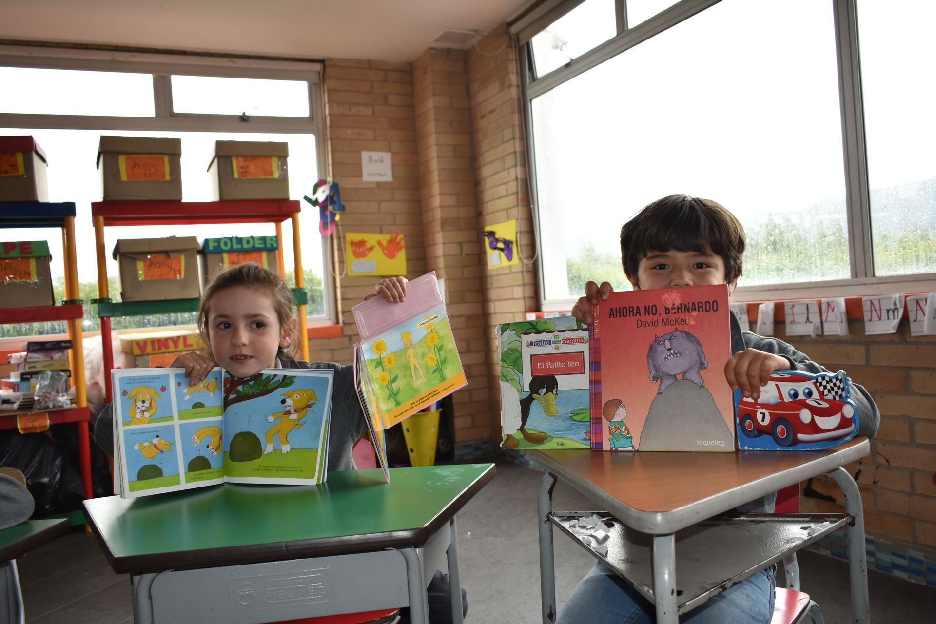 Dos niños están sentados en sus pupitres, sosteniendo coloridos libros ilustrados, en un aula luminosa.