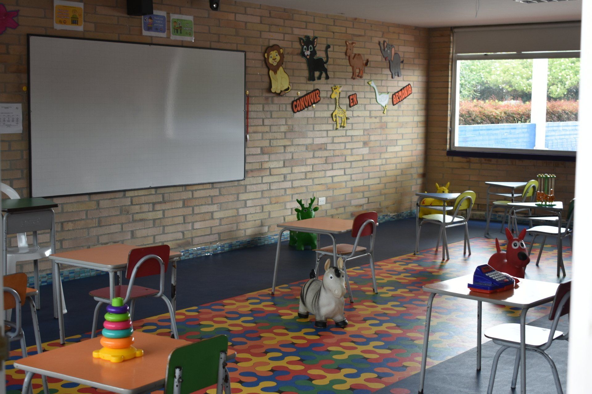 Aula vacía con pizarra blanca, alfombra colorida, sillas dispersas y una gran ventana.
