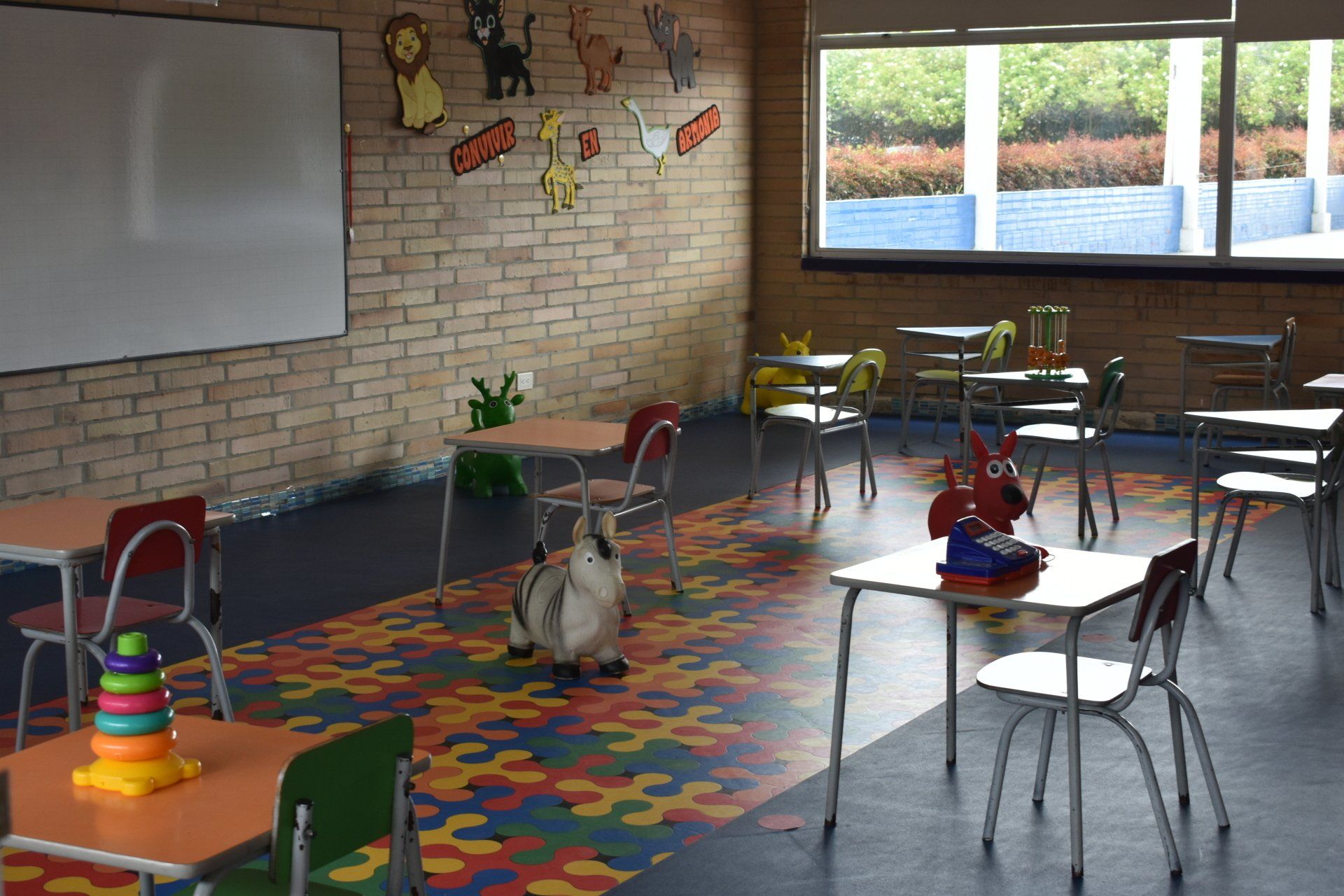 Aula vacía con alfombra colorida, pupitres, sillas, pizarra blanca y grandes ventanales junto a paredes de ladrillo.
