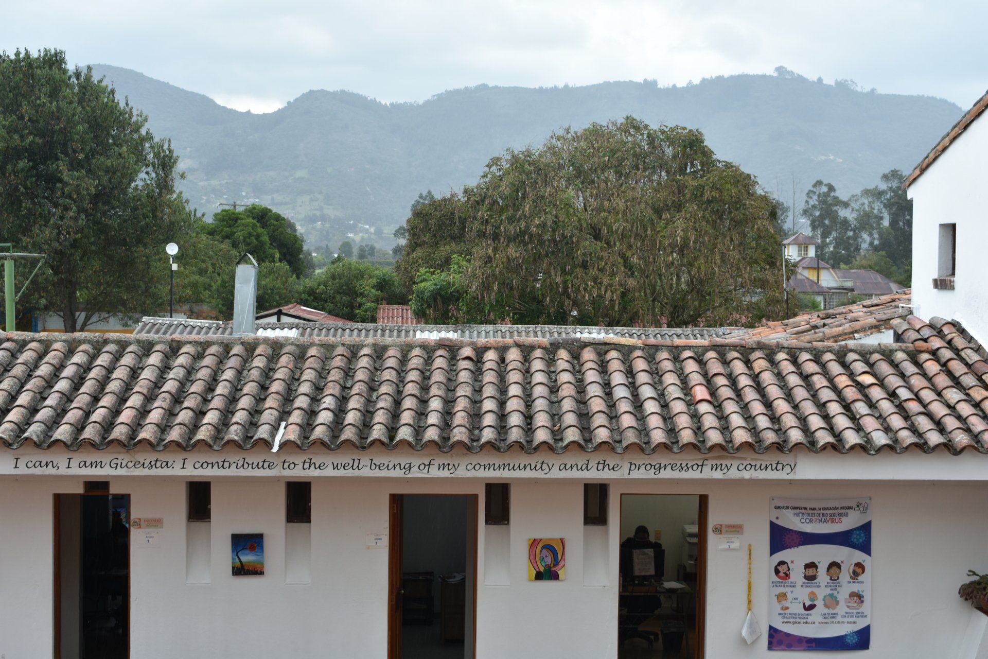 Edificio blanco con tejado de tejas en un patio, montañas y árboles al fondo.
