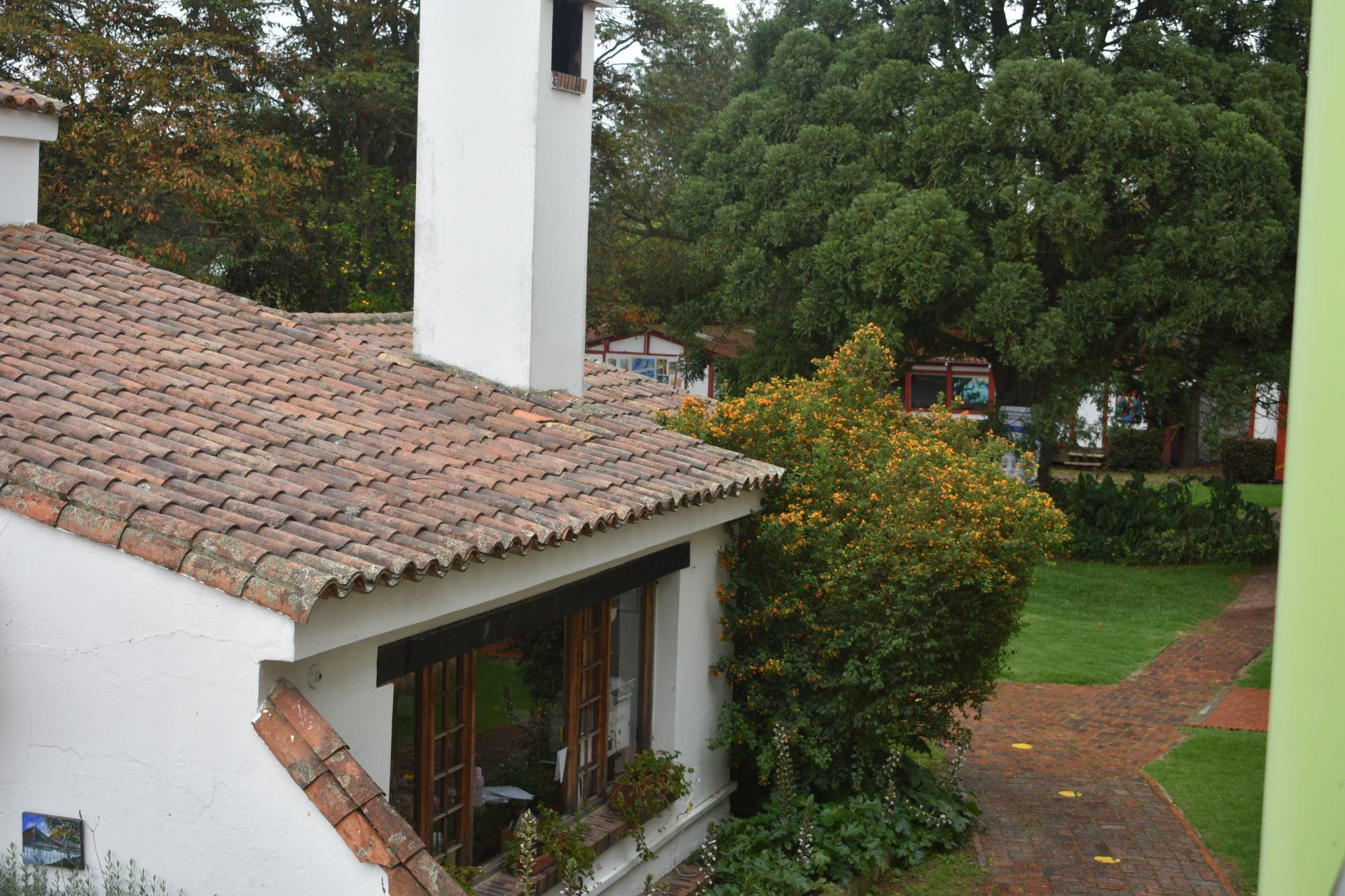 Casa blanca con tejado de tejas y chimenea junto a un jardín verde y árboles después de la lluvia.