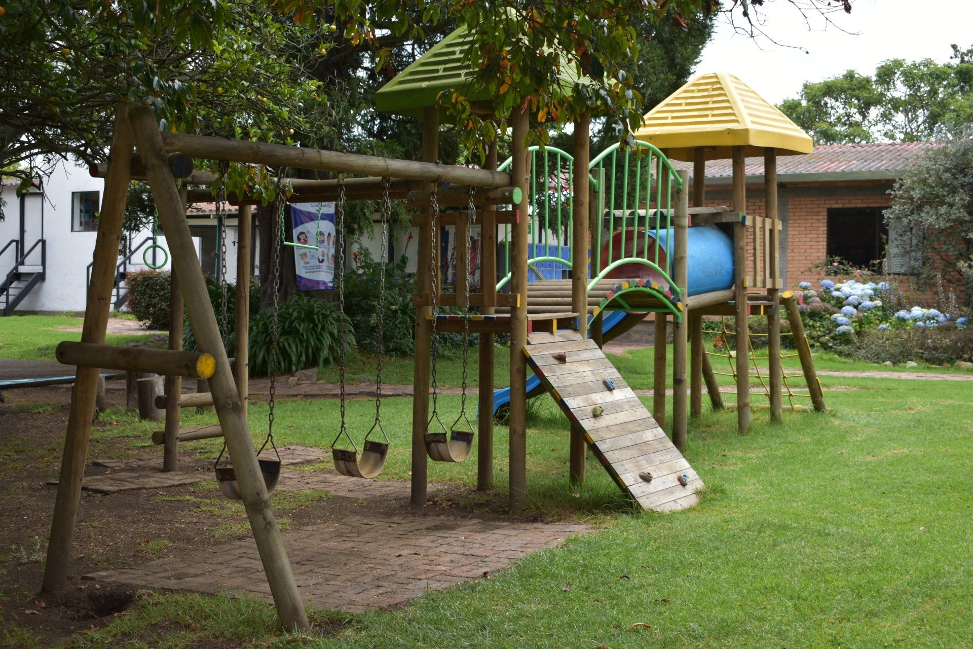 Parque infantil con estructura de madera para trepar, tobogán, columpios y zona de césped bajo los árboles.