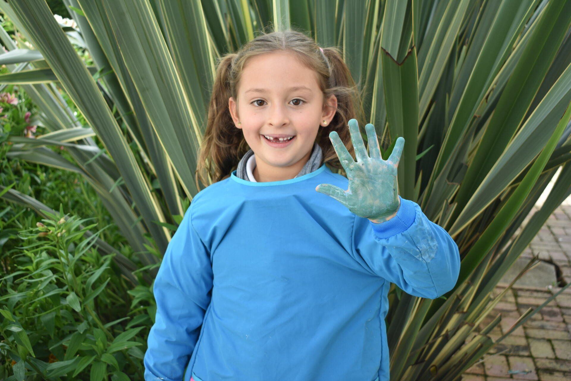 Niño con camisa azul y guantes sonriendo y mostrando una mano pintada frente a plantas verdes.