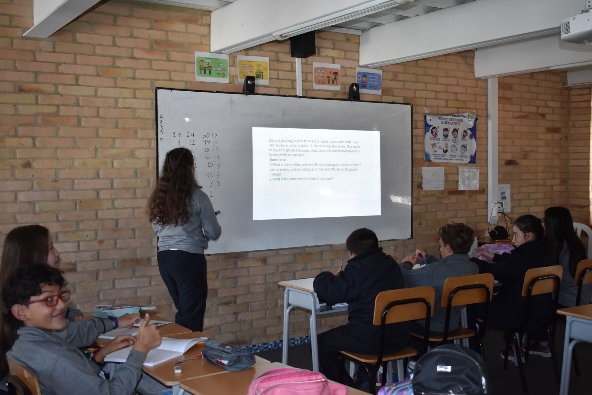 Escena en un aula con alumnos sentados mientras un profesor escribe en una pantalla proyectada al frente.