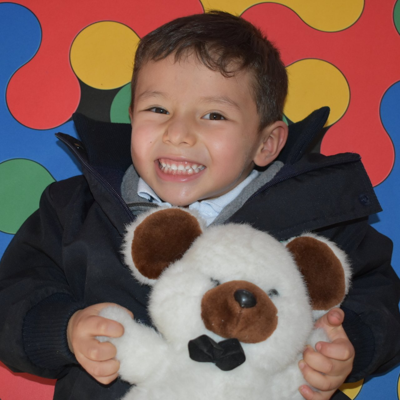 Niño sonriente sosteniendo un osito de peluche blanco sobre un fondo colorido rojo y amarillo.