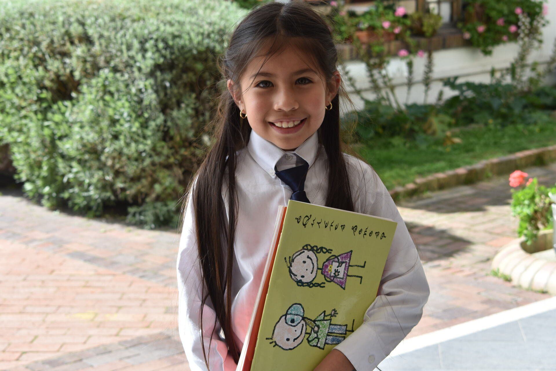 Una colegiala sonriente sostiene una carpeta de dibujos de colores al aire libre en un sendero de jardín.