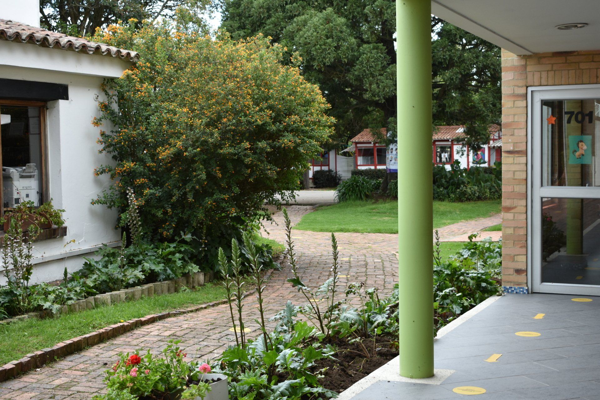 Paseo por el patio junto a edificios blancos, macizos de flores y árboles verdes, con un porche cubierto y una puerta de cristal.