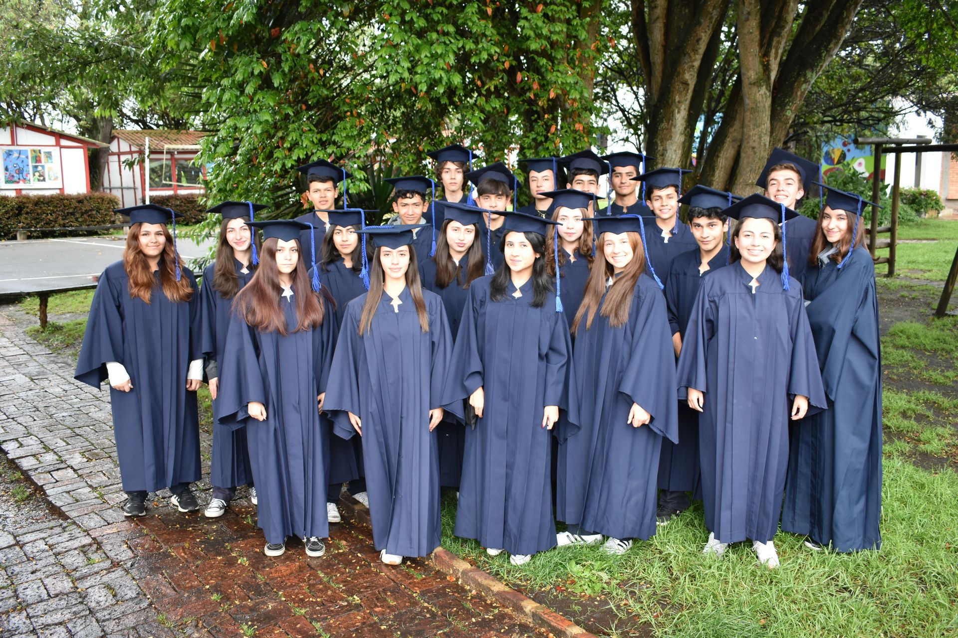 Graduados con birretes y togas azul marino posando al aire libre bajo un gran árbol.