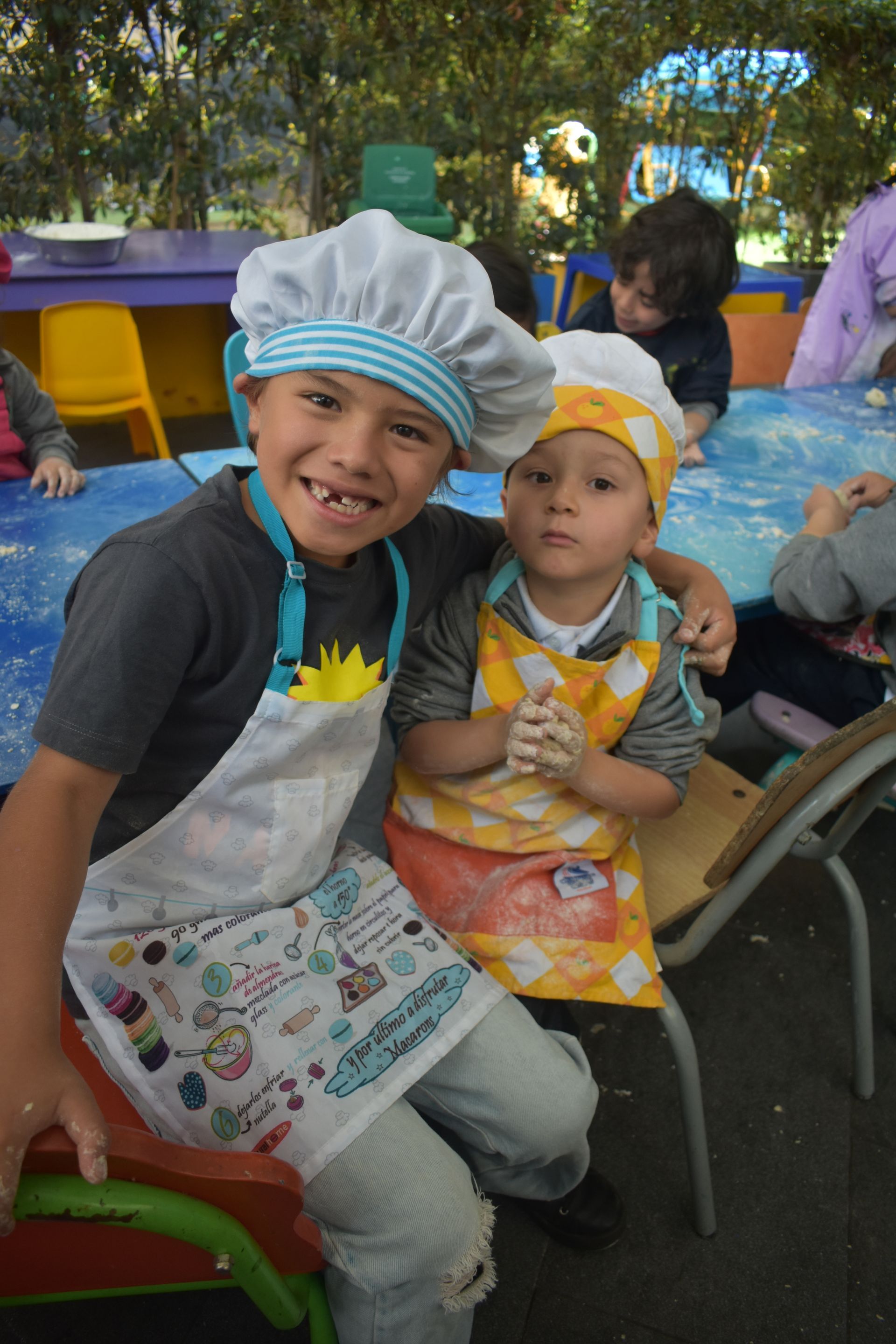 Dos niños con delantales y redecillas para el pelo están sentados en una mesa con coloridos materiales para manualidades al aire libre.