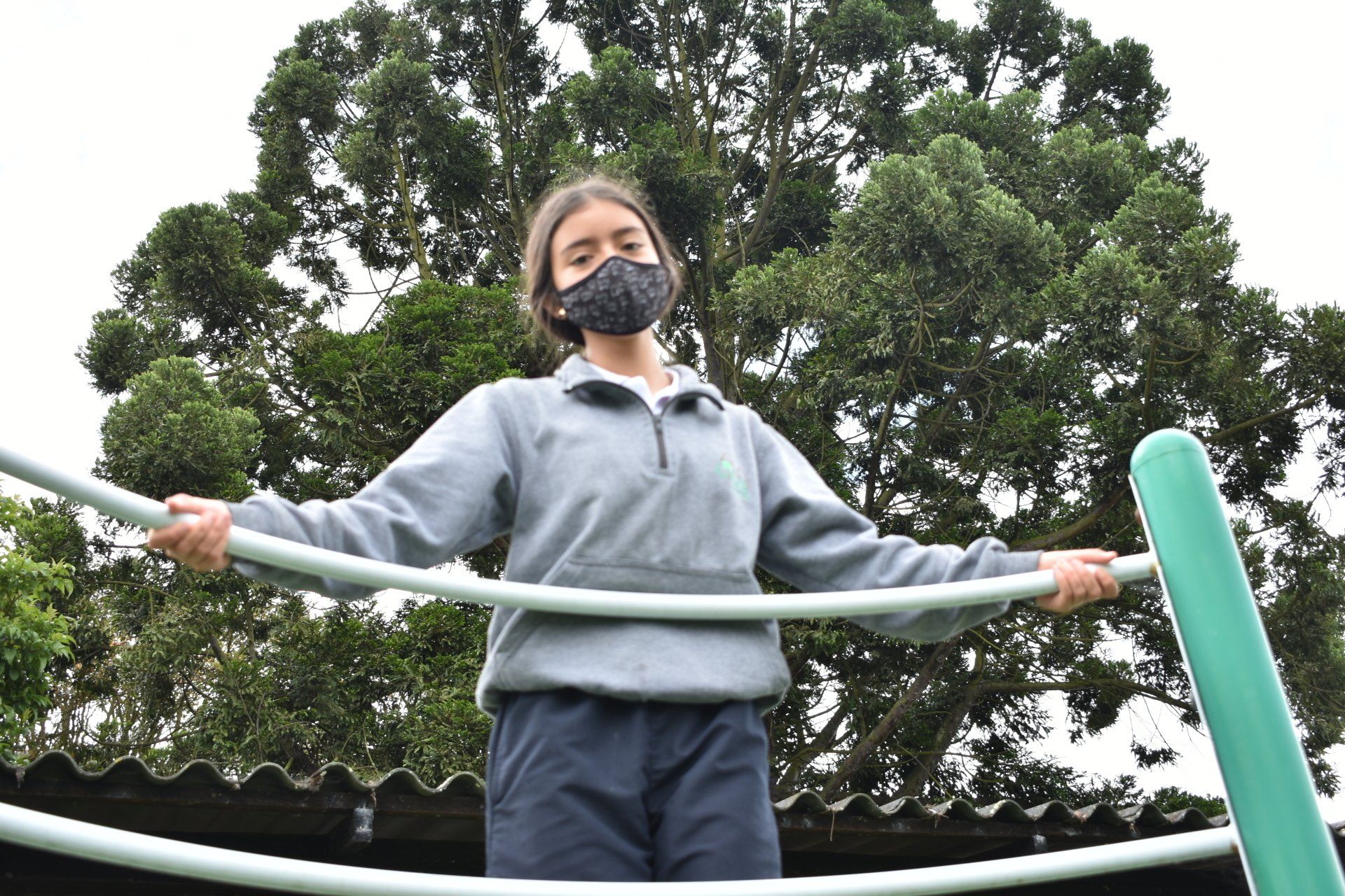 Una persona enmascarada sostiene una barra al aire libre en un parque infantil, con árboles detrás.