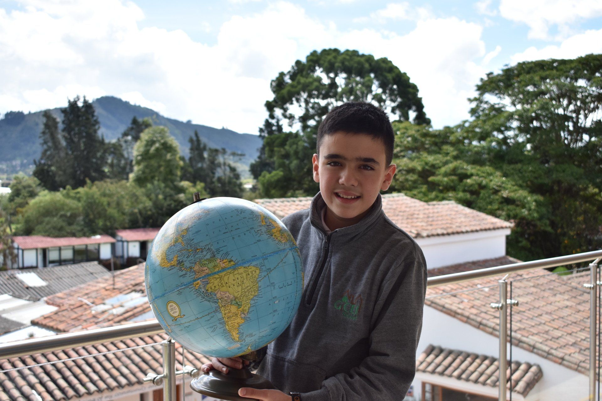 Una persona sostiene un globo terráqueo en un balcón con montañas y árboles al fondo.
